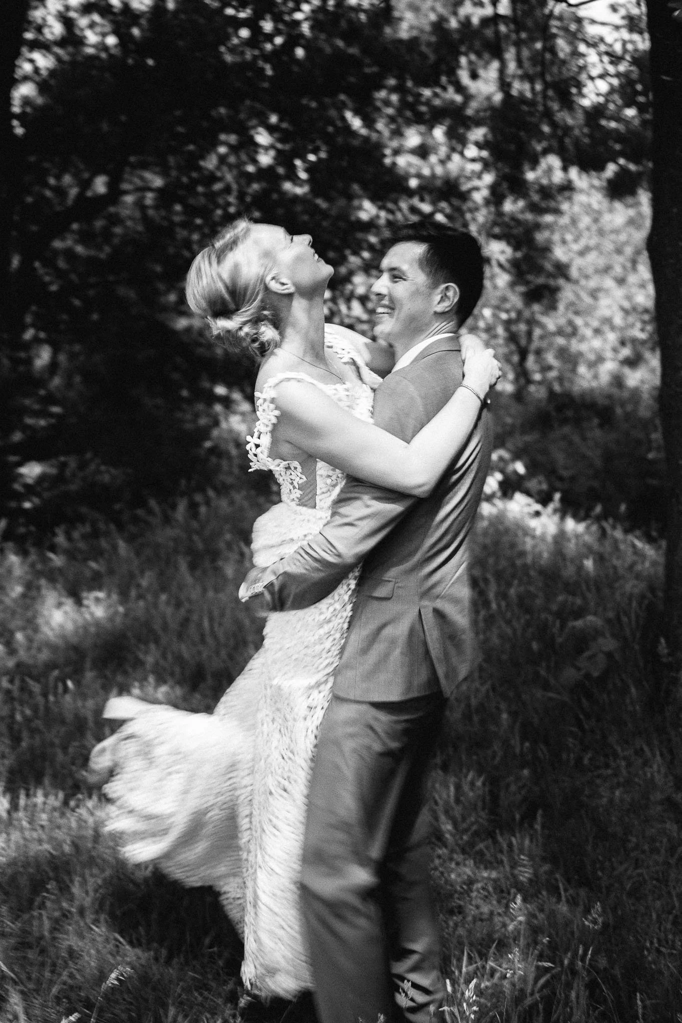Een bruid en bruidegom in een bos, de bruidegom tilt de bruid op terwijl ze lachen en dicht bij elkaar staan.