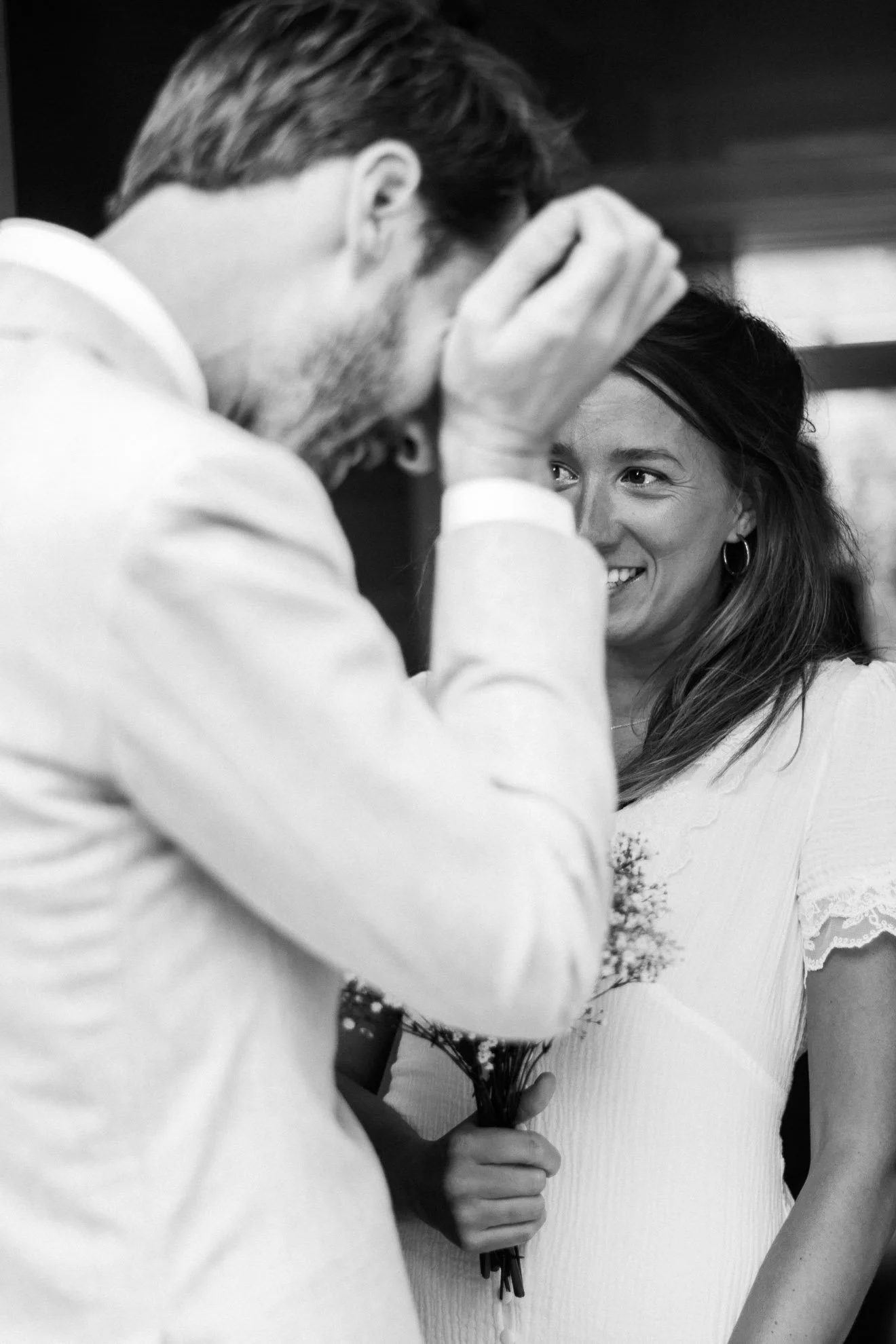 A man holding a woman's hand and touching her face gently, with the woman smiling and holding a bouquet of flowers.