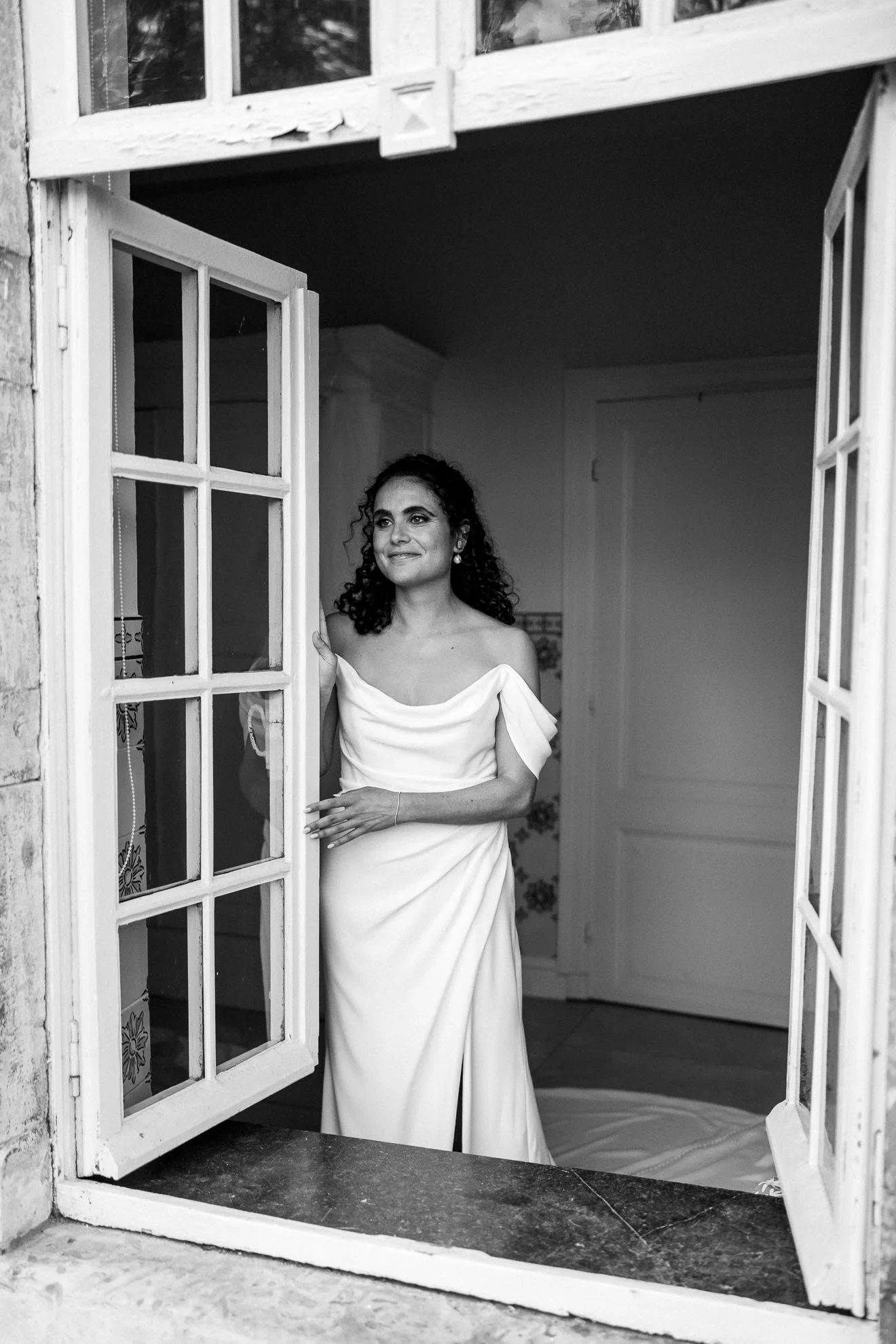 Woman in a white off-shoulder dress standing at an open window, smiling and looking outside, in a black-and-white photograph.