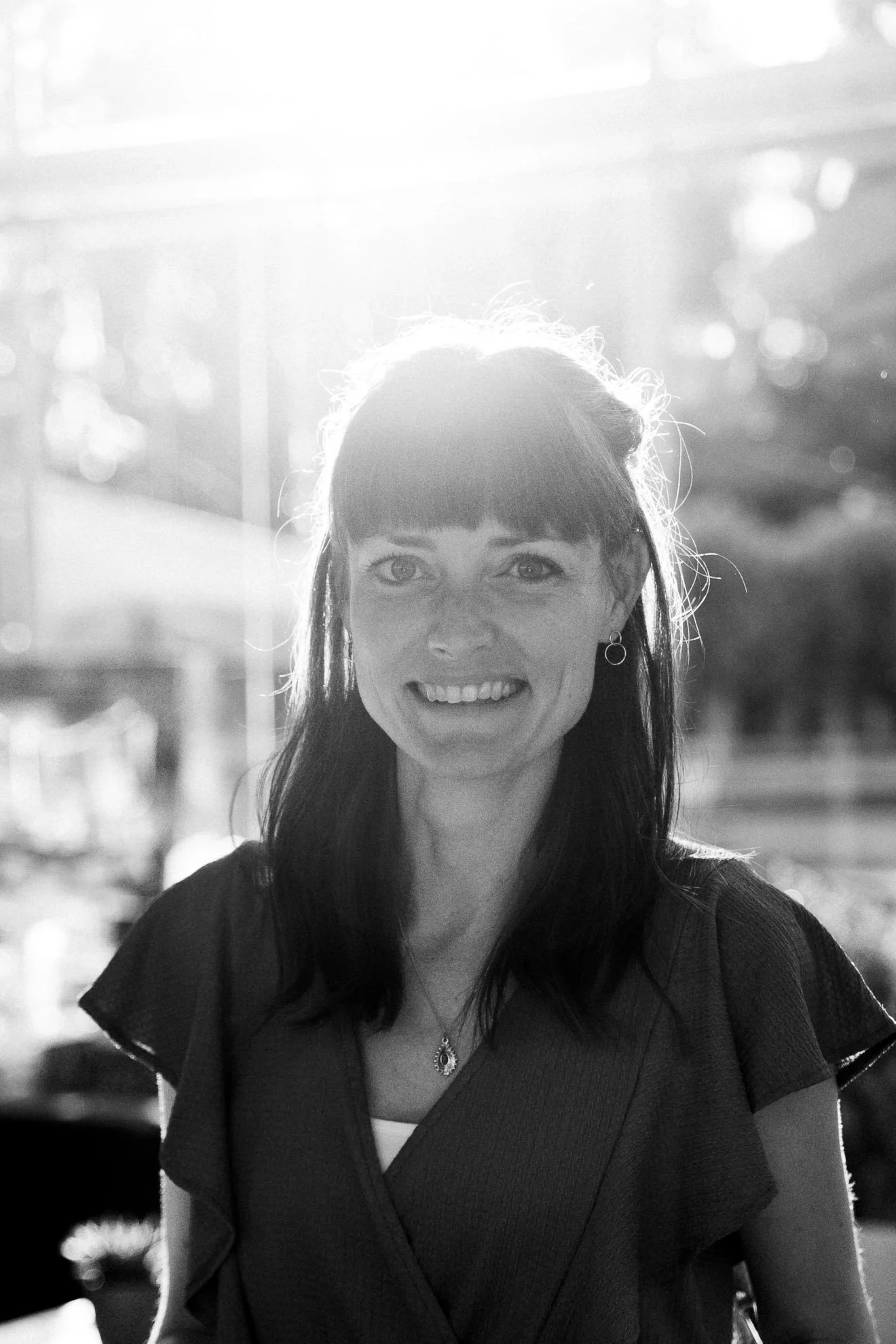 A woman with dark hair and bangs, smiling, in black and white, with sunlight creating a halo effect around her head. She is wearing earrings and a necklace, and is sitting indoors with a blurred background.