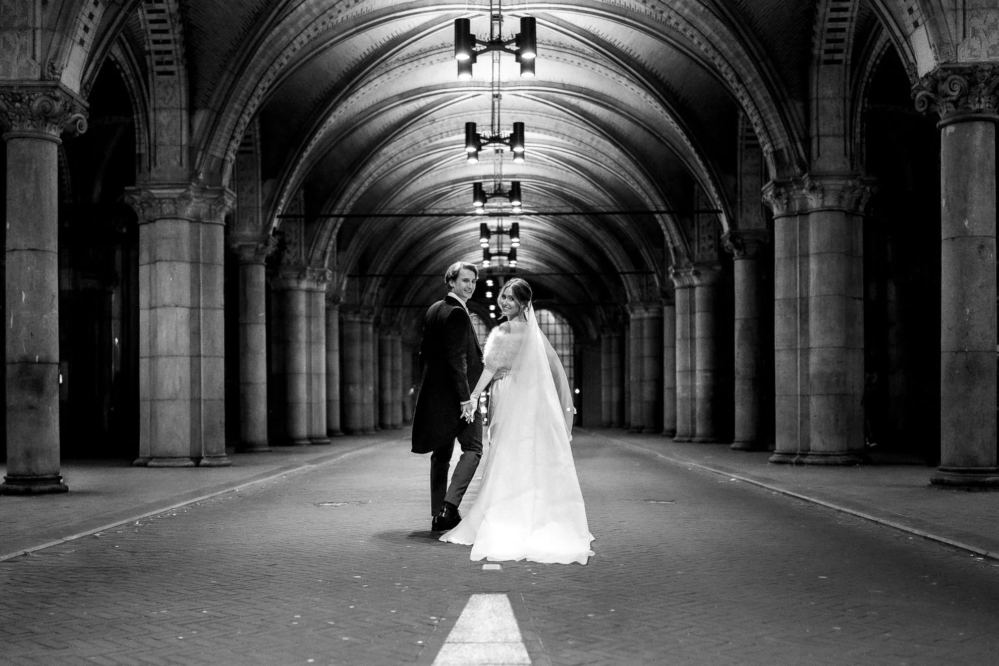 A couple in wedding attire holding hands and smiling in an underground tunnel with arched ceilings and columns, lit by overhead lights in black and white.
