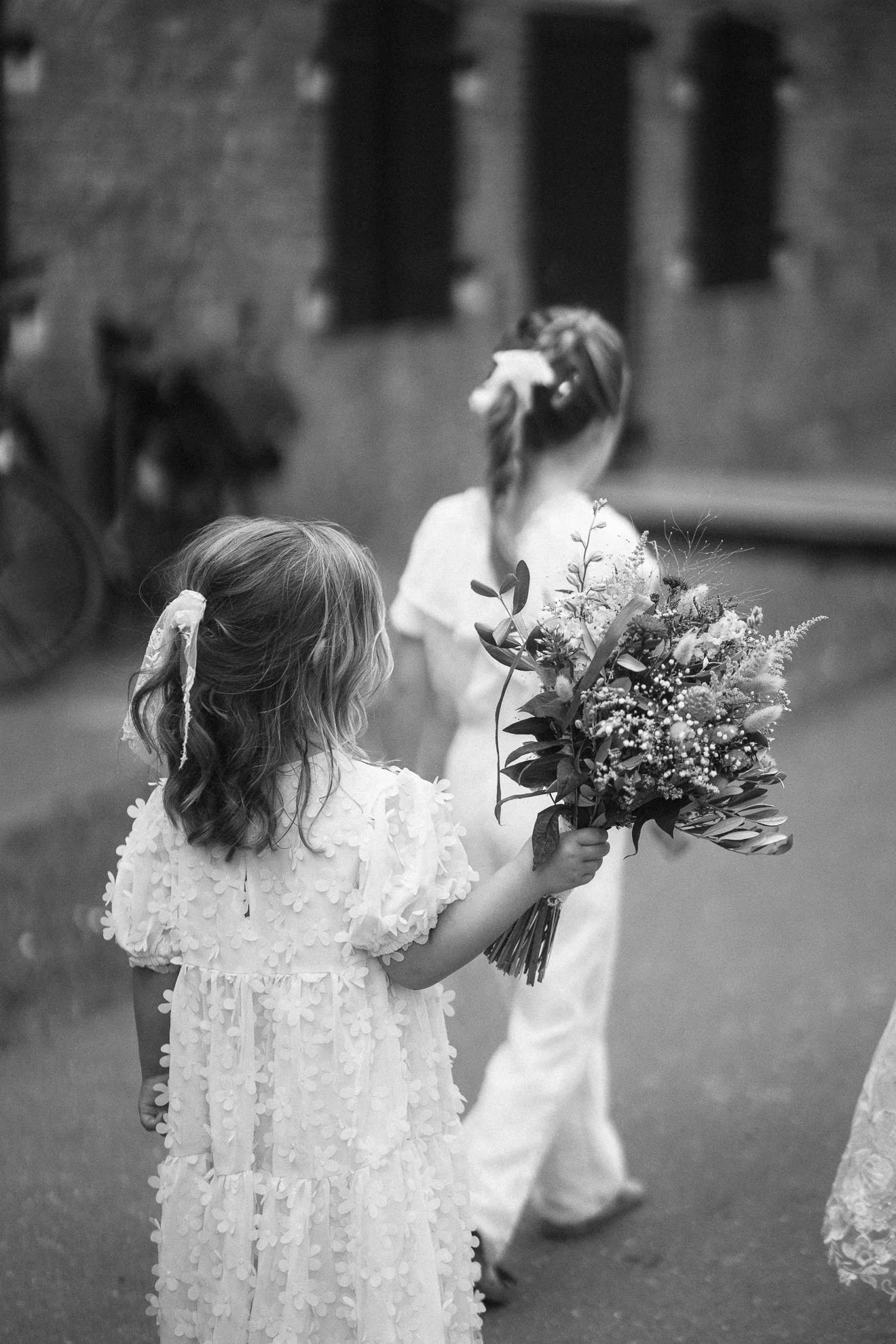 A young girl in a white dress holding a bouquet of flowers at a formal outdoor event, with other children in similar dresses and a brick building in the background.