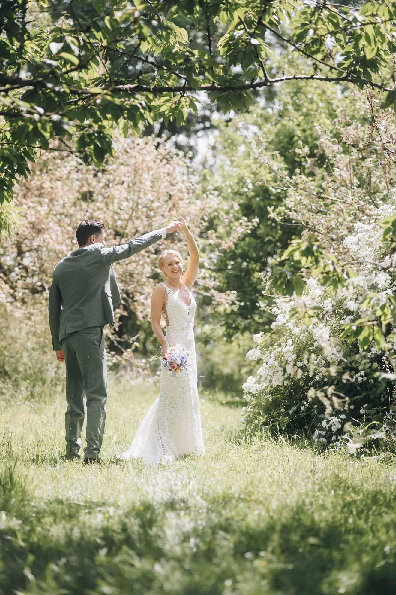 A bride and groom dancing outdoors among blooming trees, with the groom twirling the bride who is holding a bouquet, in a sunlit garden setting.