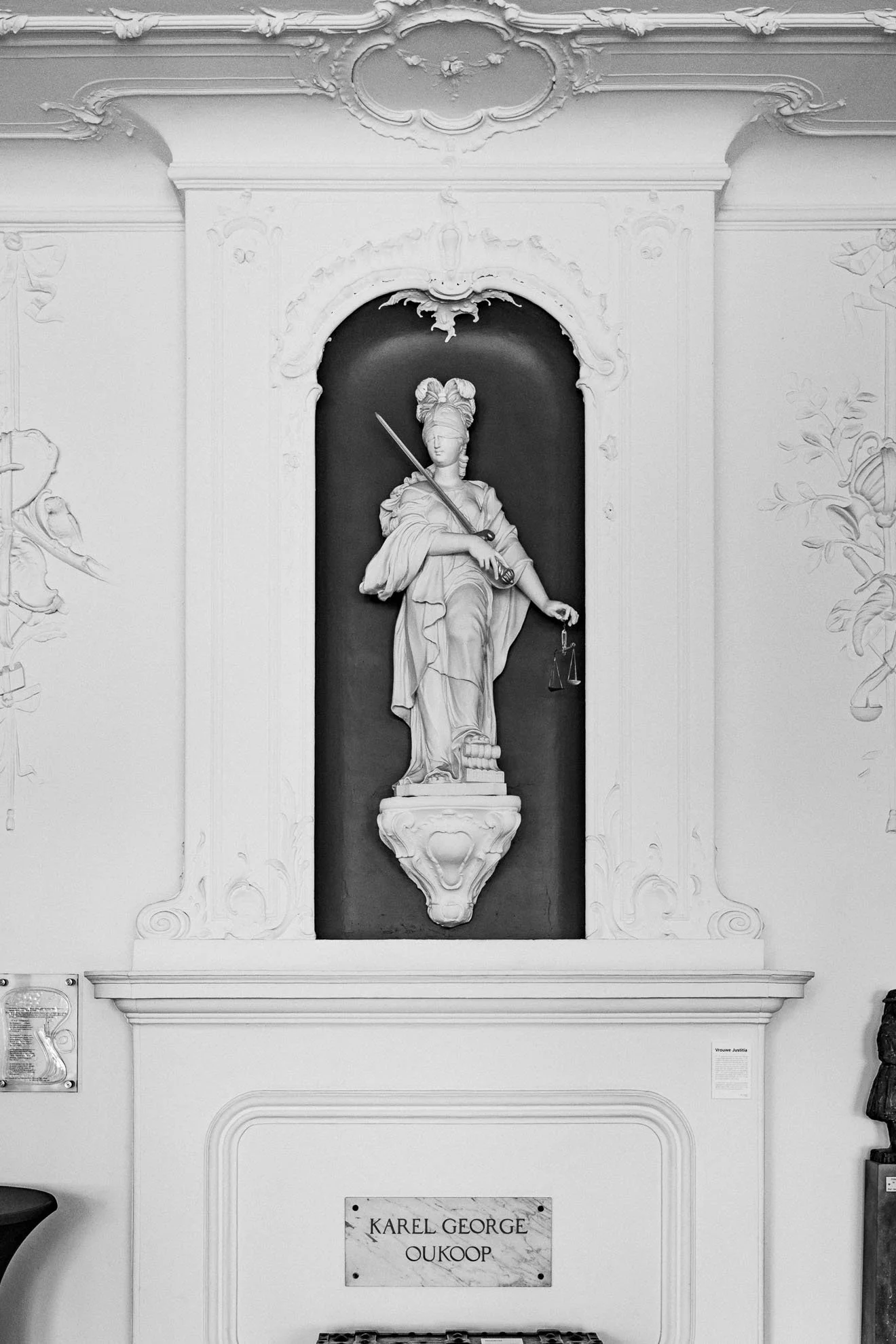 A white sculpture of a woman in classical attire holding a spear and scales, set in an ornate alcove with decorative molding. A plaque below reads 'Karel George Oukoop'.