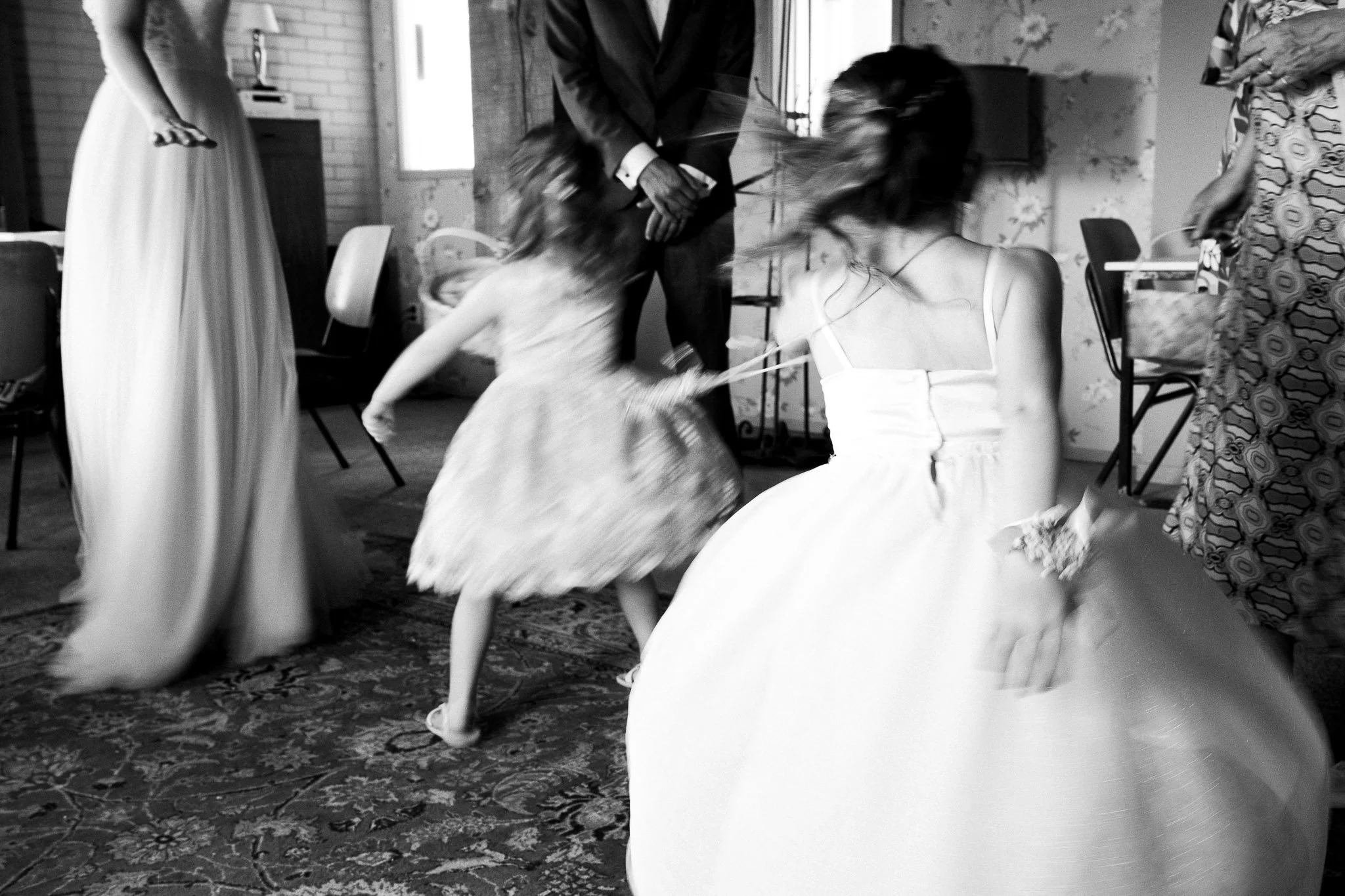 Een zwart-wit foto van een groep mensen op een feest of bruiloft, met een jonge meid in een feestjurk die lijkt te dansen of te spelen, terwijl twee andere vrouwen ook in jurk staan.