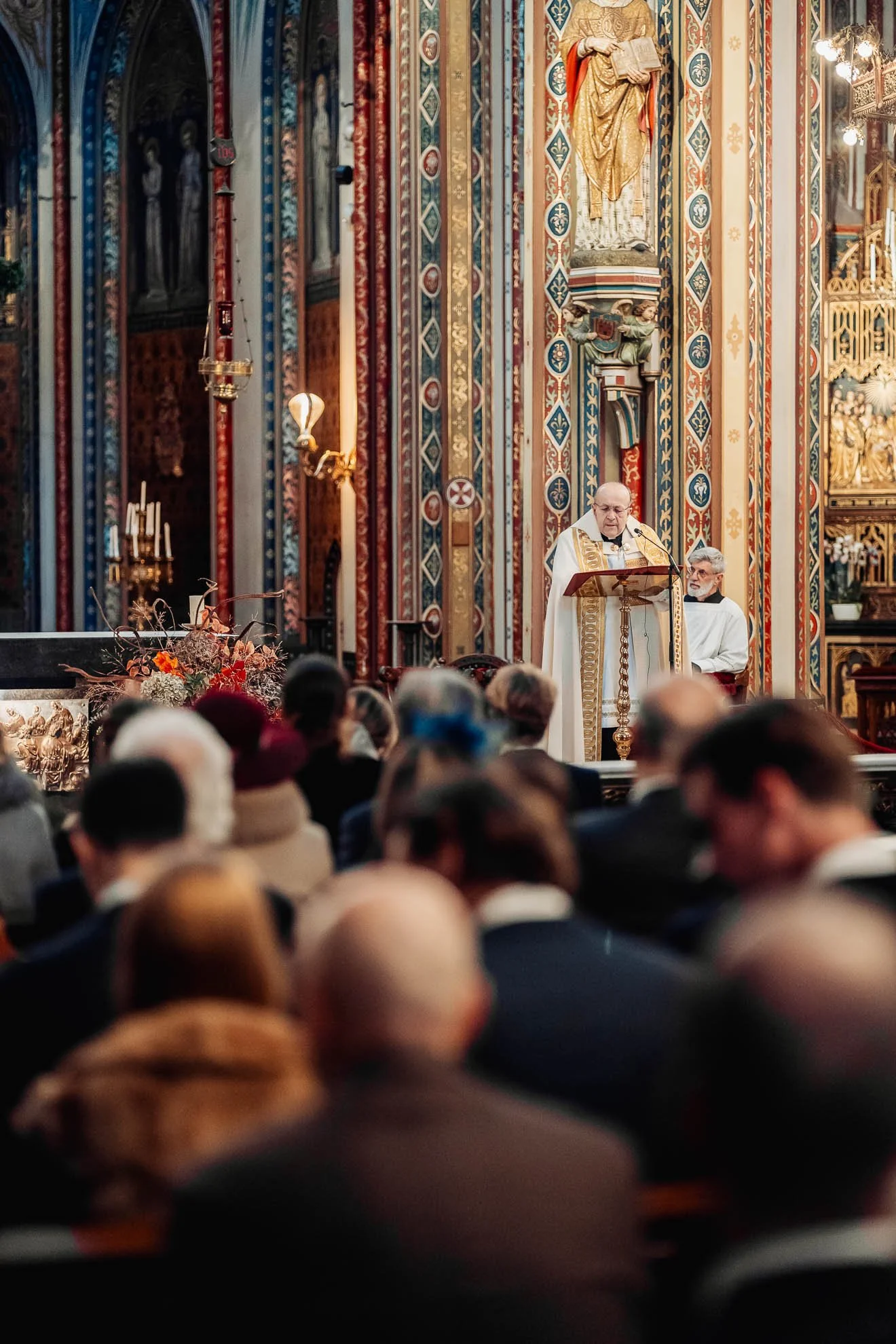 A religious ceremony inside a decorated church with ornate golden and colorful artwork. A priest is standing at the pulpit reading from a book, while another clergy member sits nearby. The congregation is seated and listening. The church interior fea
