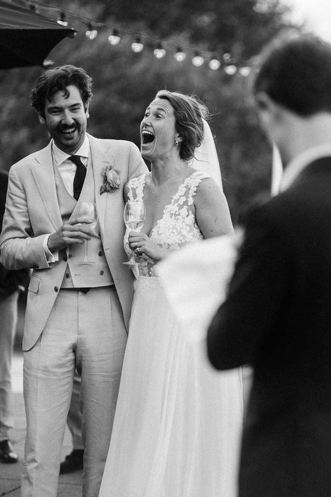Een zwart-wit foto van een bruid en bruidegom die lachen en elkaar vasthouden tijdens een bruiloft, met gasten op de achtergrond.
