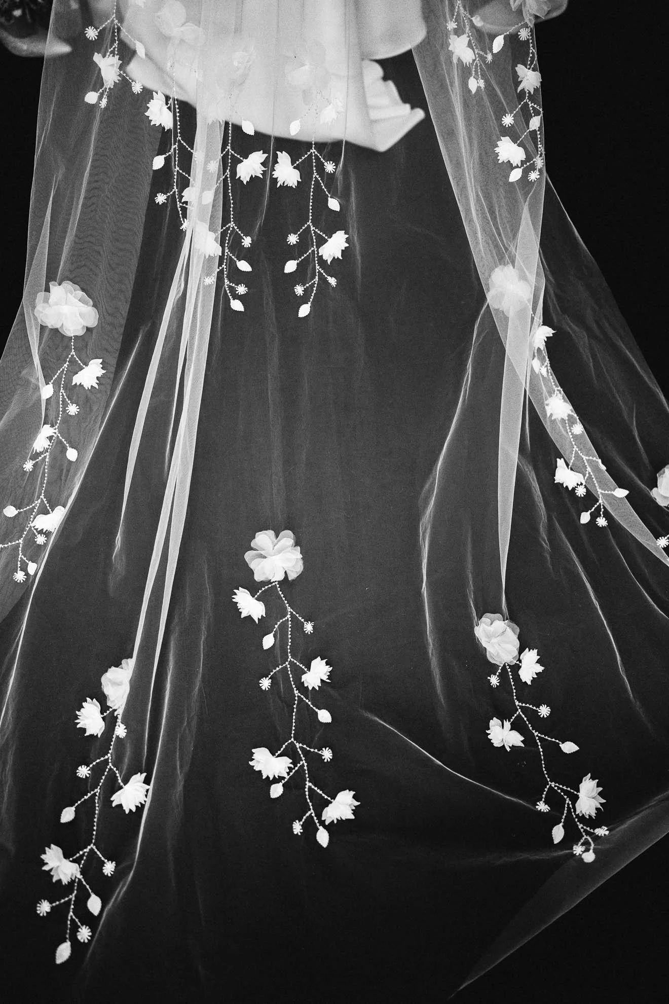 Close-up of decorative sheer fabric with white floral and leaf embroidery, hanging against a dark background.