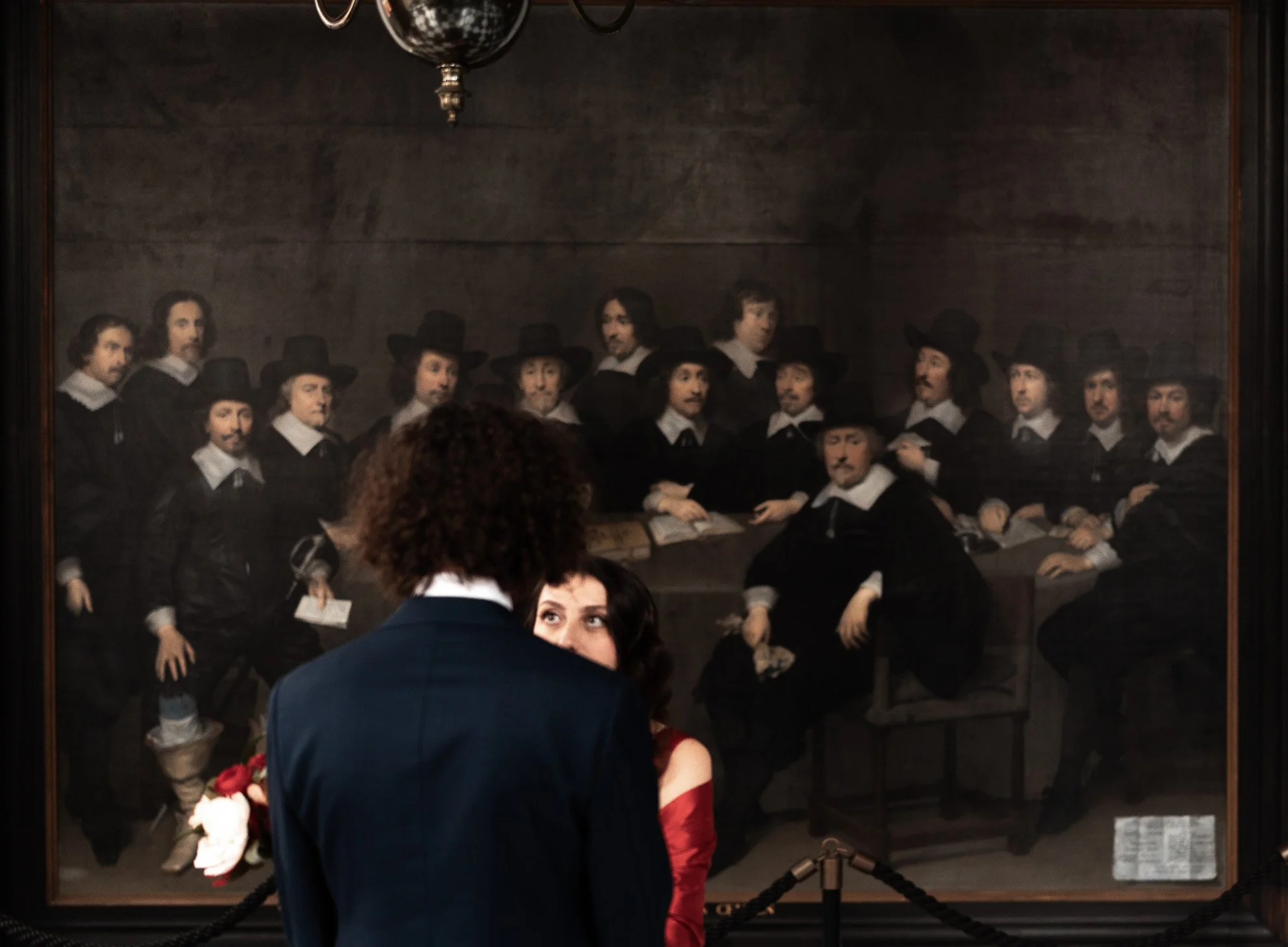 People viewing a famous painting of a group of men in historical clothing, with a chandelier above and dark wood wall background.