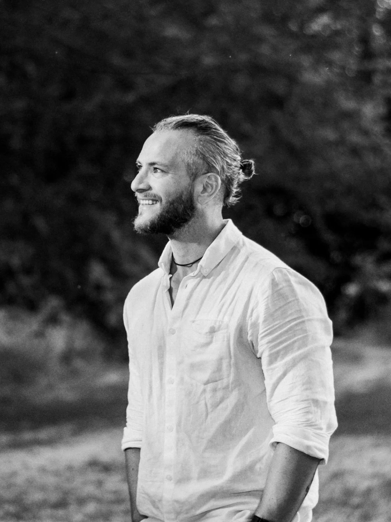 Een man met een baard en gespierd gezicht, die glimlacht en naar links kijkt, in een witte blouse, buiten tegen een achtergrond van bomen en struiken.