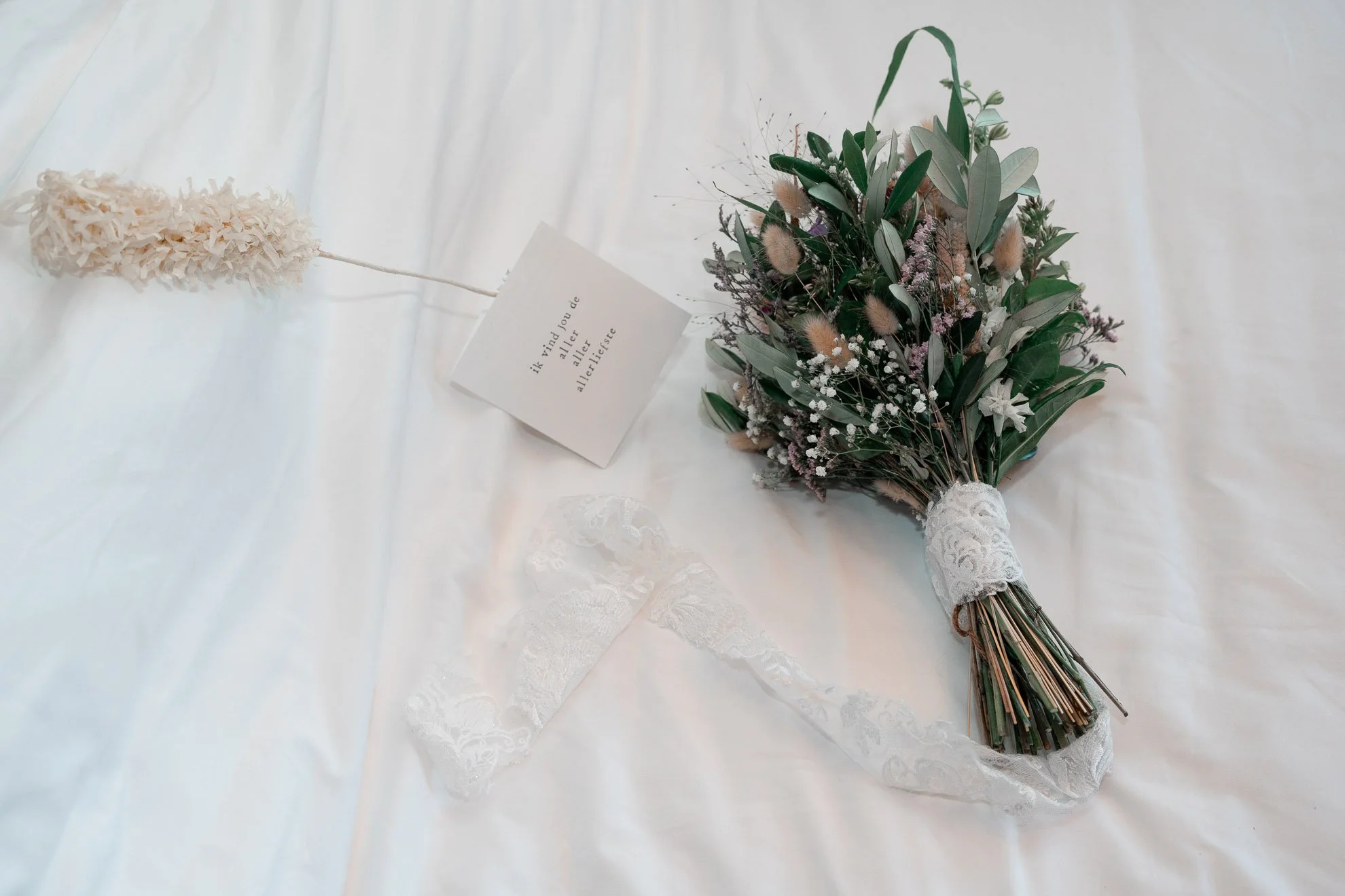 A bouquet of greenery and assorted flowers lying on a white fabric surface. The bouquet is wrapped at the base with lace, and there is a white card with text in a different language nearby. There is also a fluffy, cream-colored decorative object atta