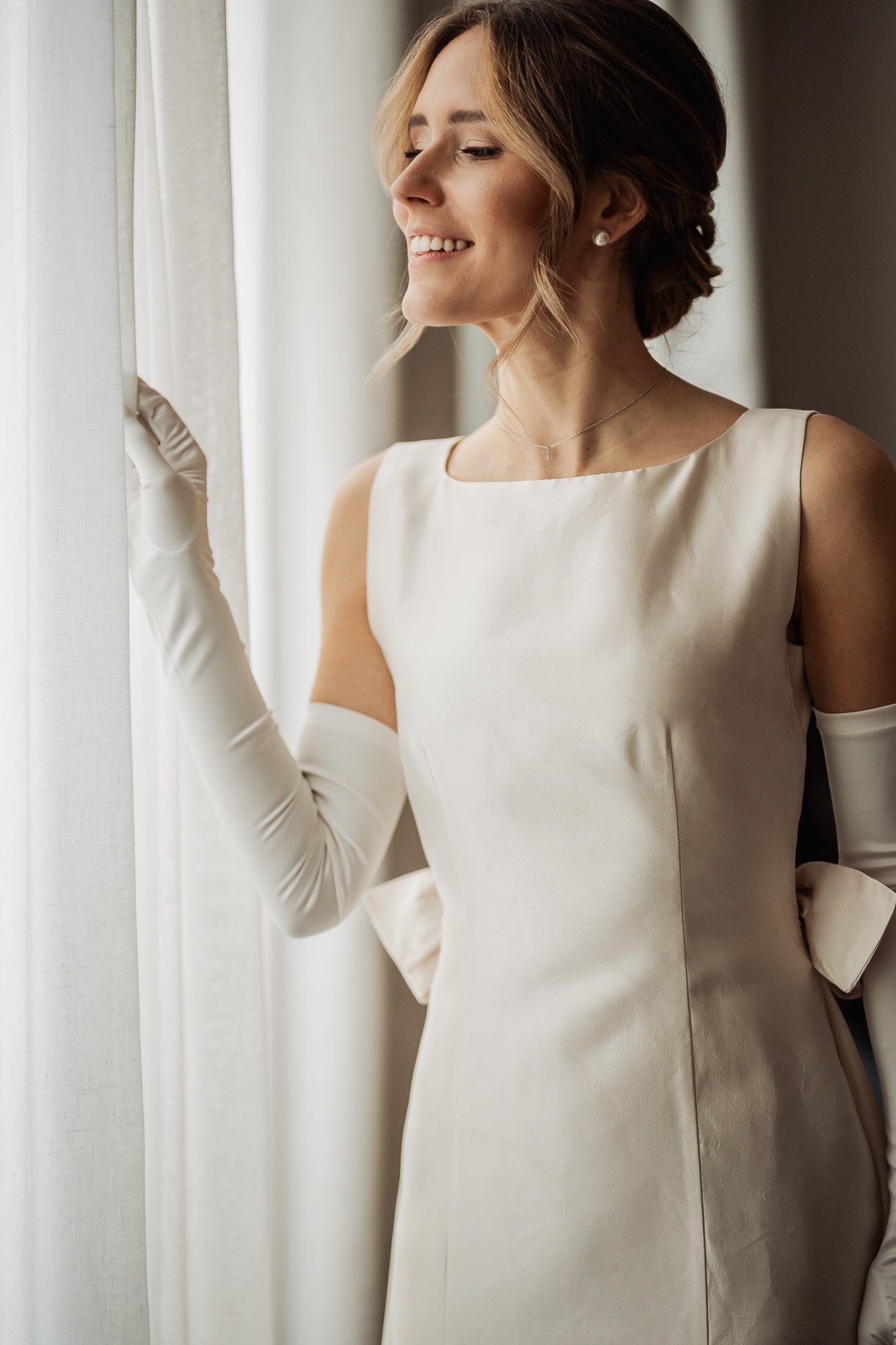 A woman with shoulder-length hair wearing a white elegant dress with satin gloves, standing near a window with sheer white curtains.