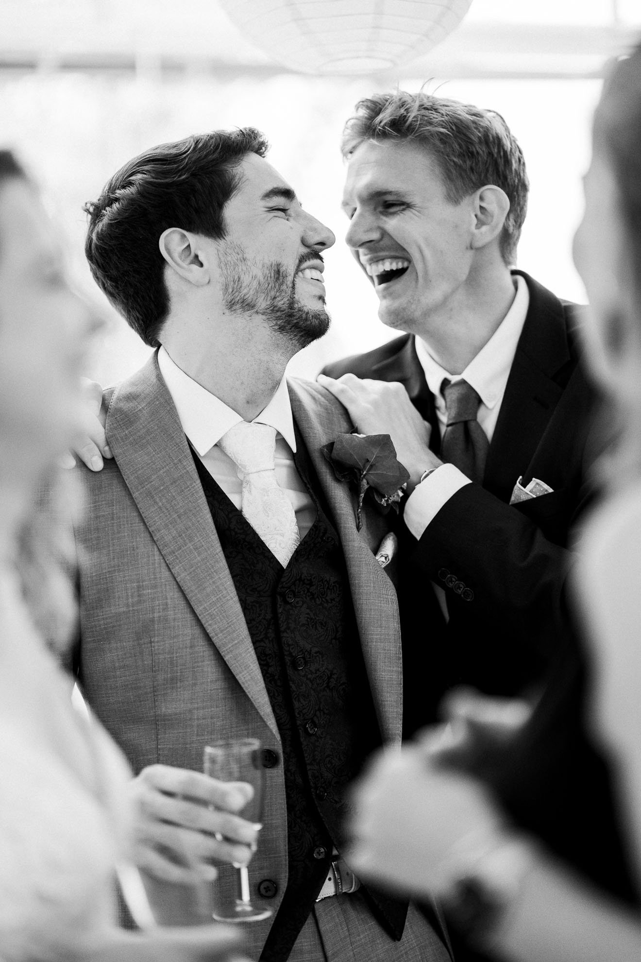 Black and white photo of two men smiling at each other at a social gathering, with blurred people in the background. One man is holding a drink.