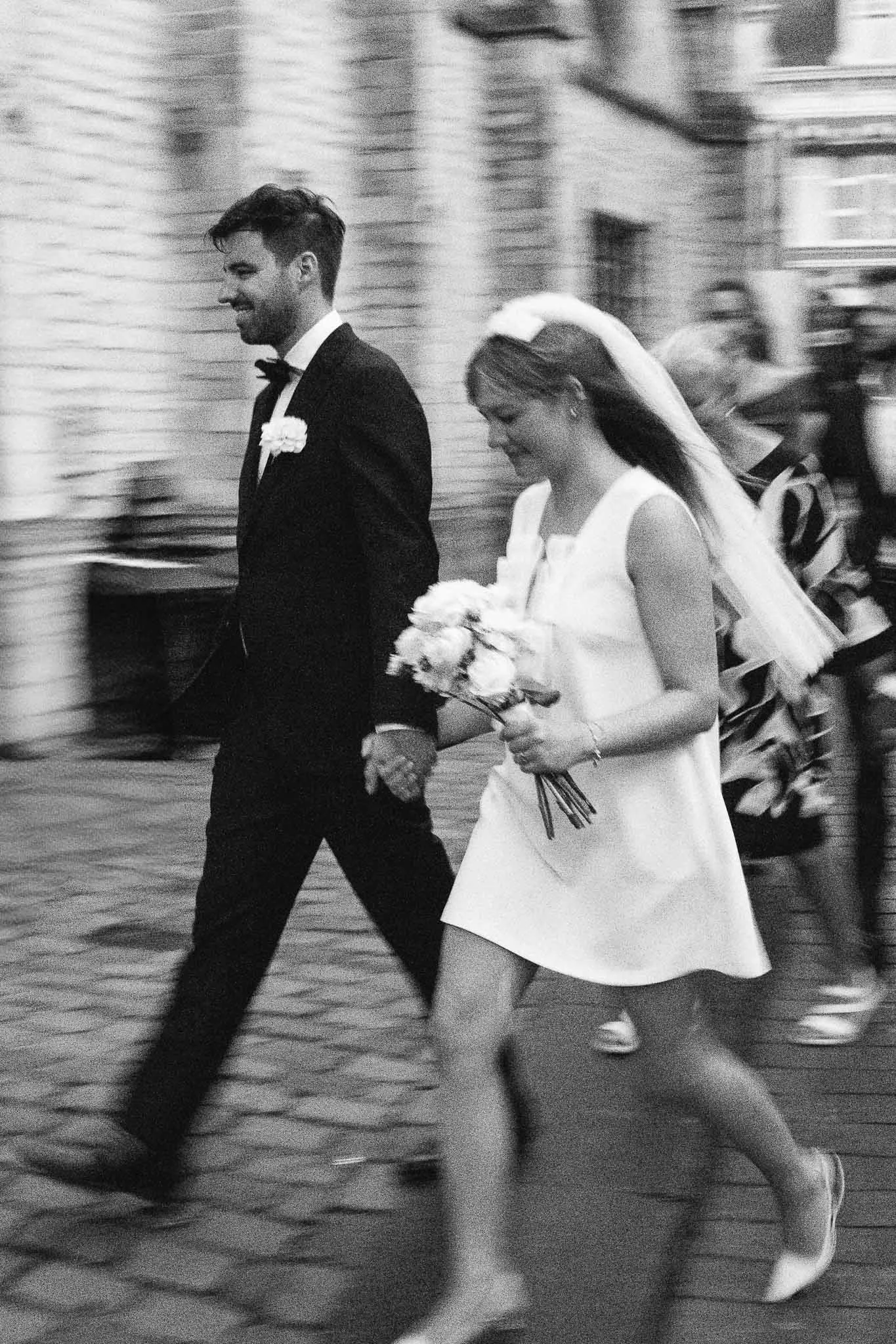 een zwart-wit foto van een getrouwde paar die samen lopen, de man in een zwart pak en de vrouw in een witte jurk met bloemen, ze houden elkaars handen vast en dragen een trouwboeket.
