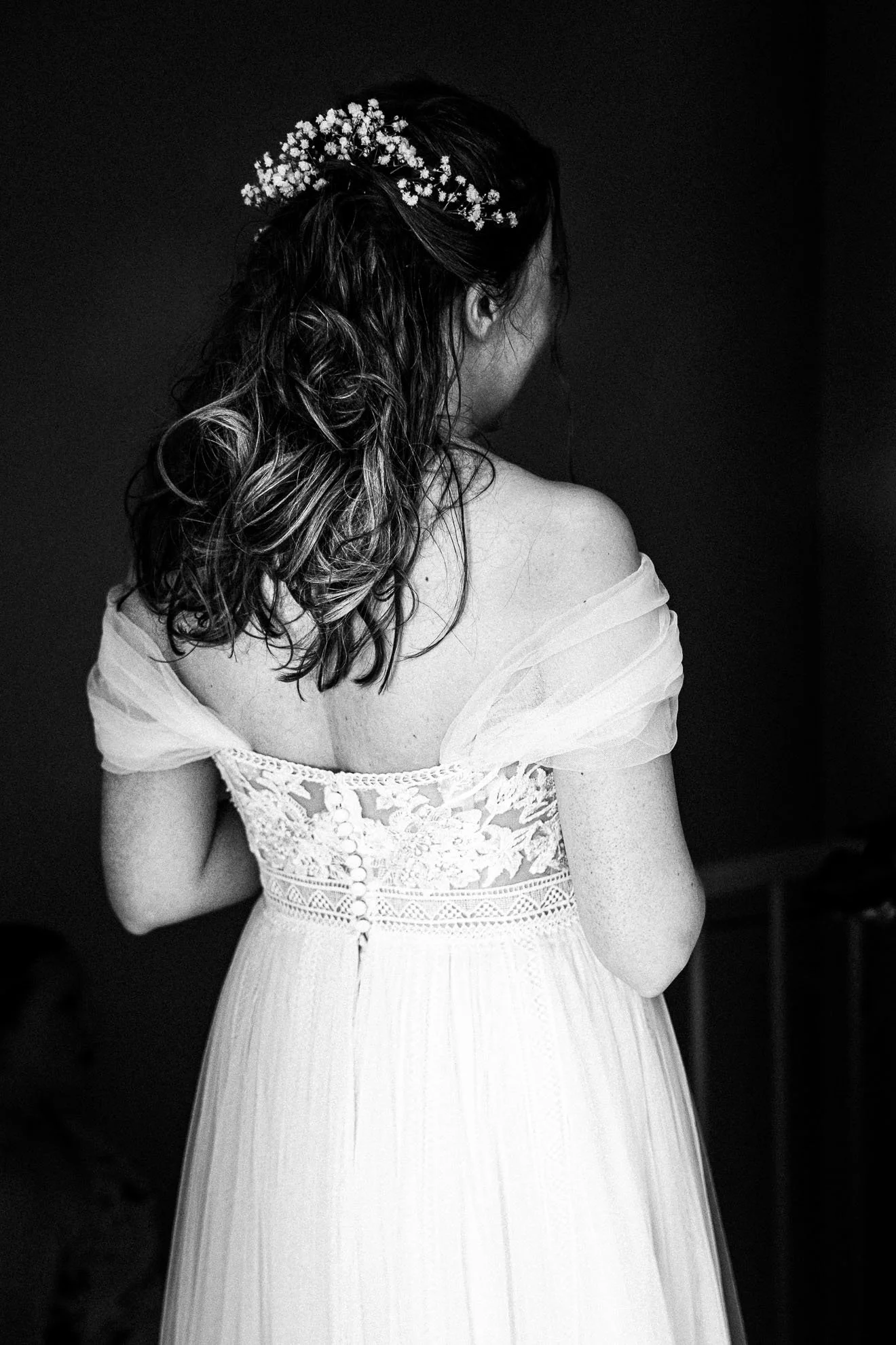 Back view of a woman in a wedding dress with lace details, wearing a floral hair accessory, against a dark background.