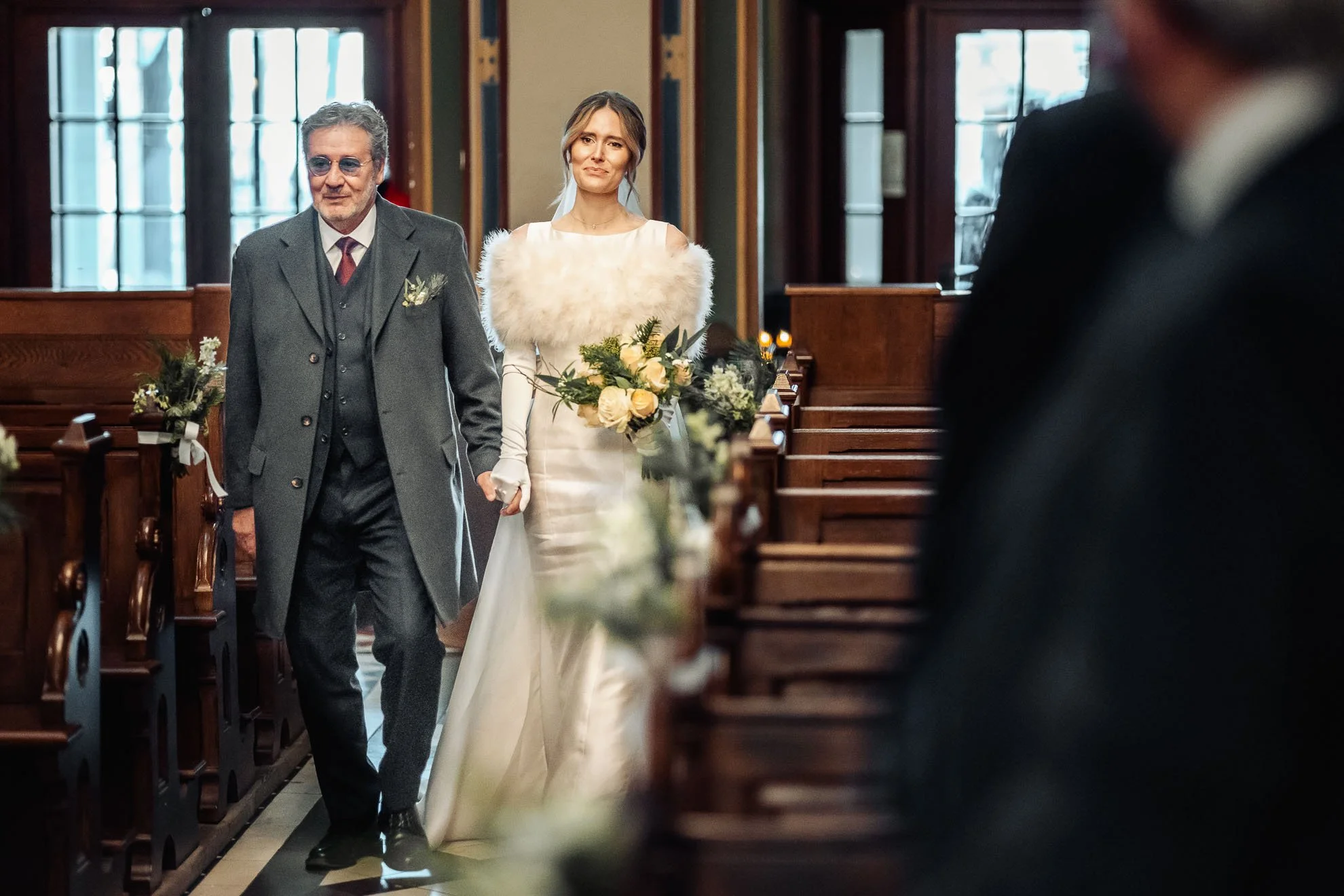 A bride in a white wedding dress holding a bouquet walking down the aisle with a man in a gray suit at a wedding ceremony inside a church.