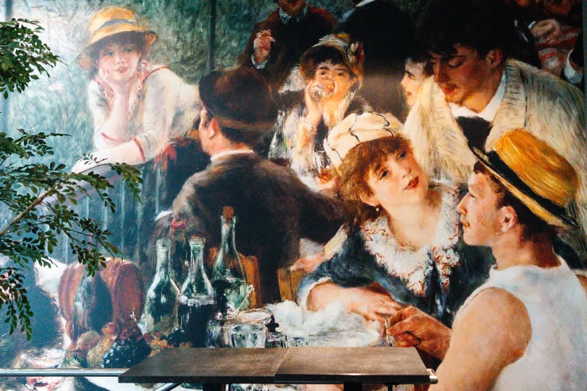 A group of people gathered around a table with drinks, engaged in conversation, next to a large, colorful Impressionist-style painting of women and children at a fountain, with greenery in the foreground.