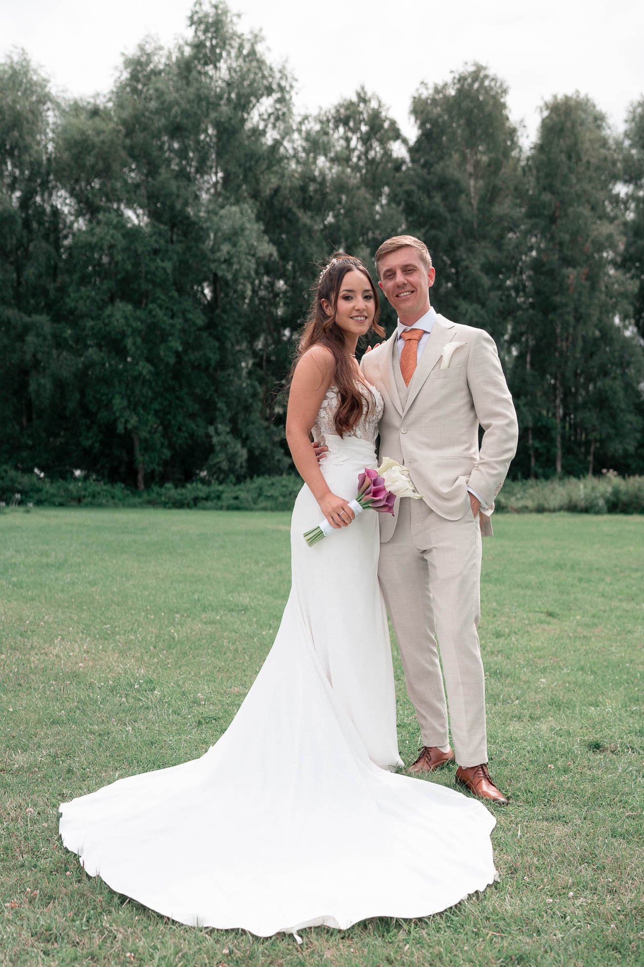 A bride and groom standing on a grassy field with trees in the background, smiling for a photo. The bride is wearing a white wedding dress and holding a bouquet of calla lilies. The groom is dressed in a beige suit with a white shirt and an orange ti