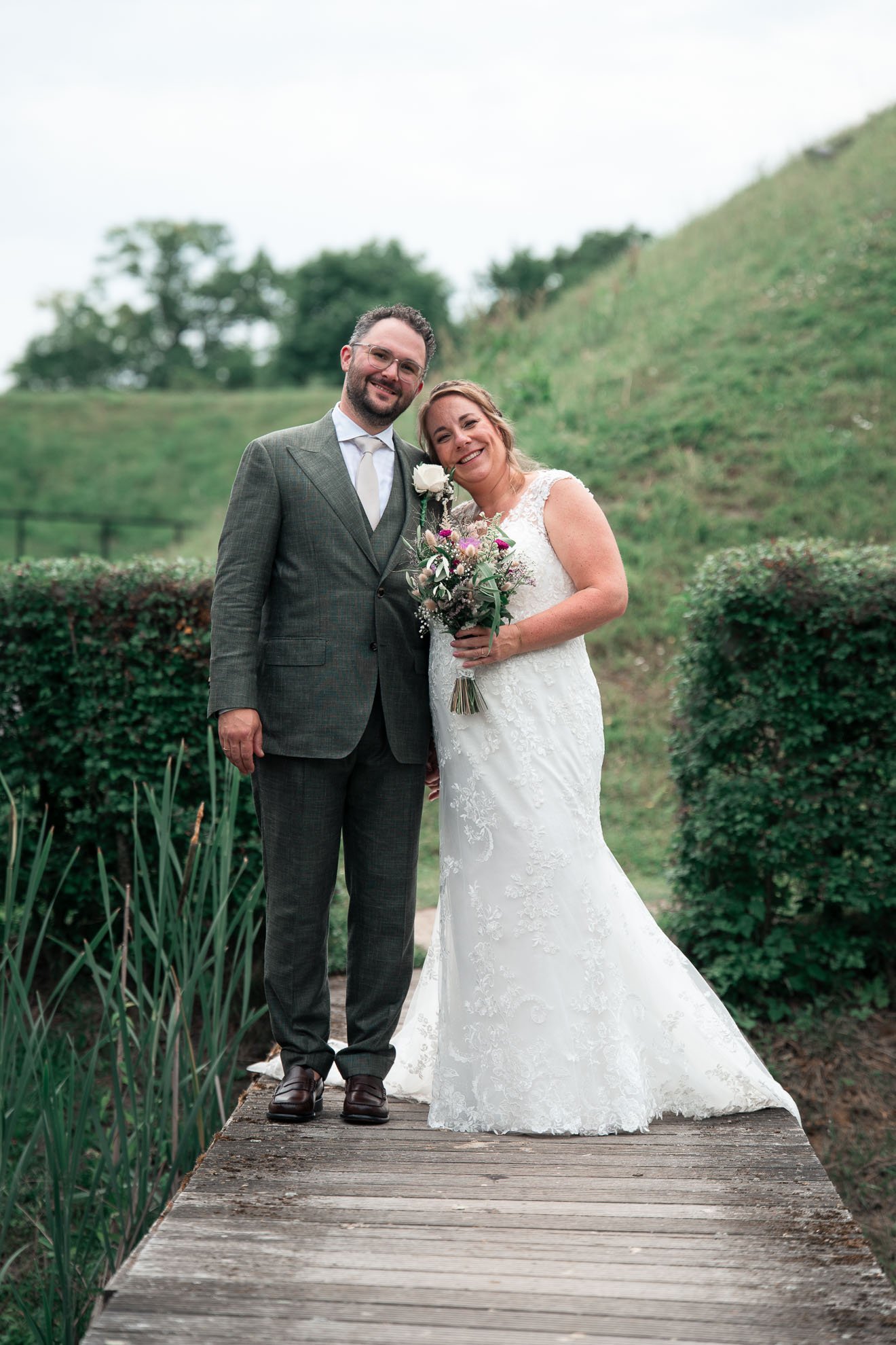 A bride and groom stand close together on a wooden bridge outdoors, smiling. The bride wears a white lace wedding dress and holds a bouquet, while the groom wears a gray patterned suit with a white shirt and tie. The background features green bushes 