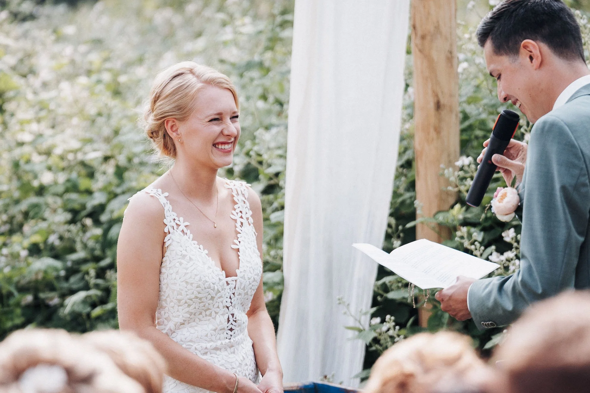 A bride with blonde hair in a floral lace wedding dress smiling during her wedding ceremony, standing opposite a groom in a light gray suit holding a microphone and reading vows, outdoors with green foliage in the background.