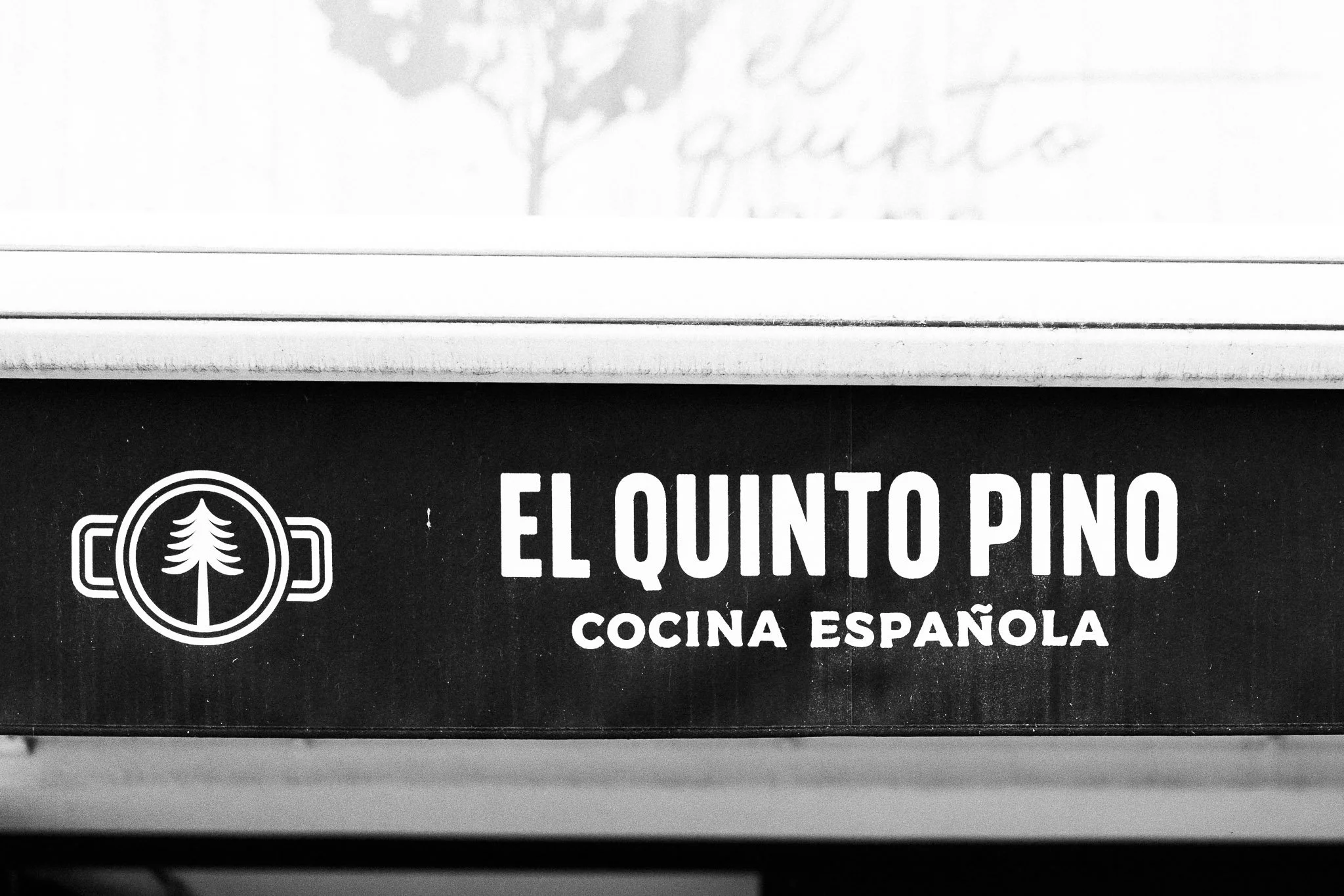 Afbeelding van een zwart-wit bord met de tekst 'El Quinto Pino' en 'Cocina Española', met een logo van een dennenboom binnen een cirkel.