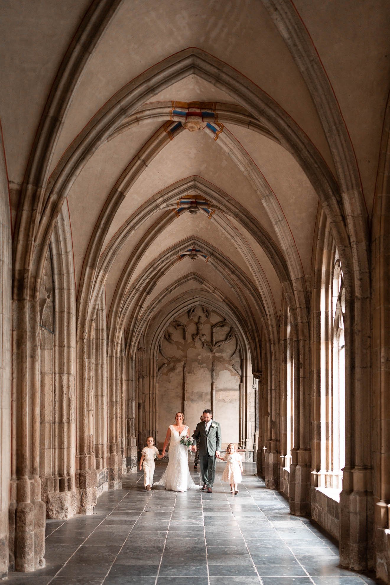 A bride and groom walking in a grand, Gothic-style cathedral under a vaulted ceiling, accompanied by two flower girls, one on each side.