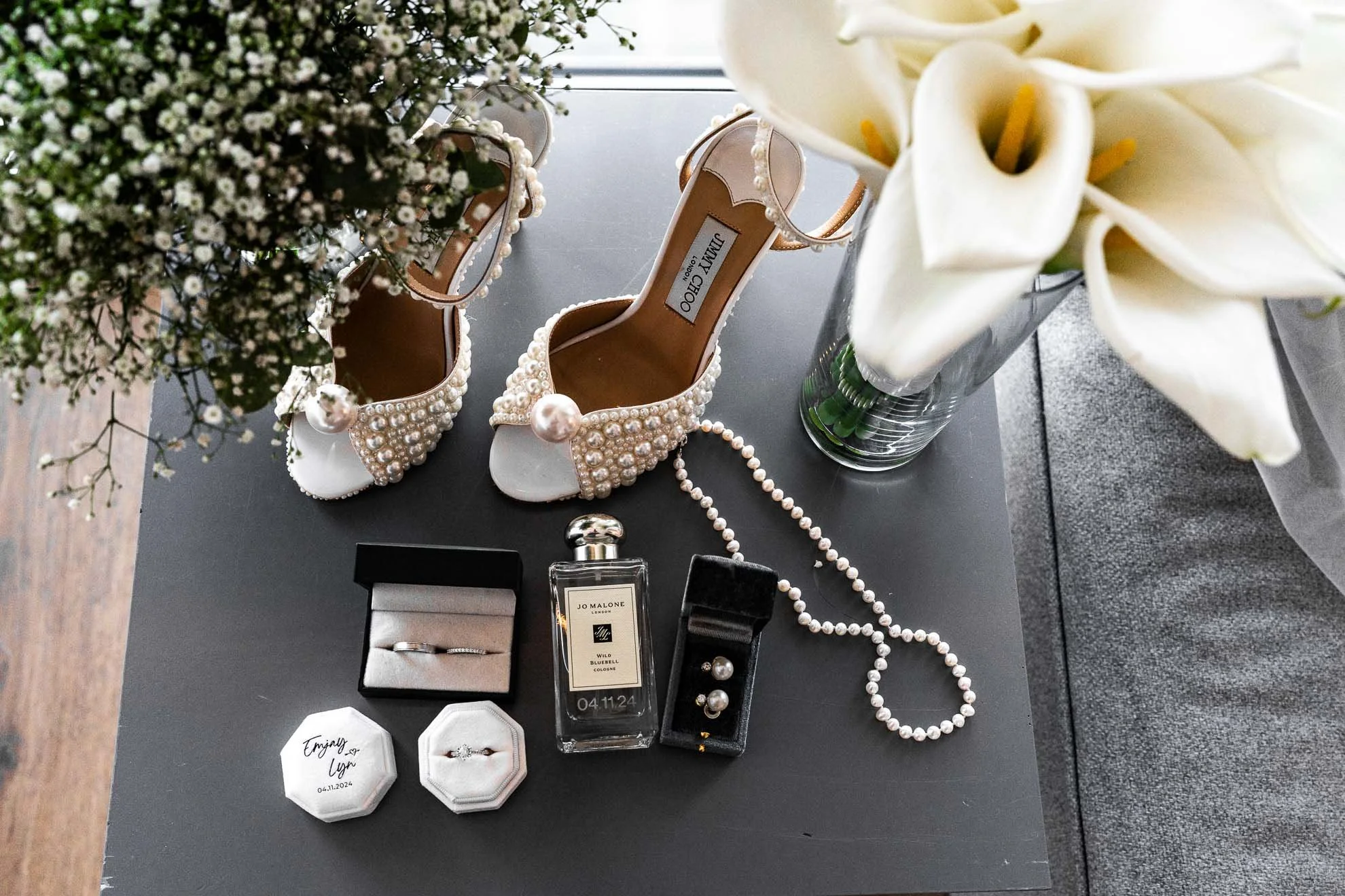 A flat lay of wedding accessories including pearl-embellished high heels, a pearl necklace, a pair of pearl earrings, a white lily flower in a glass vase, bottles of perfume, a black jewelry box with pearl earrings, and a white ring box with a ring w