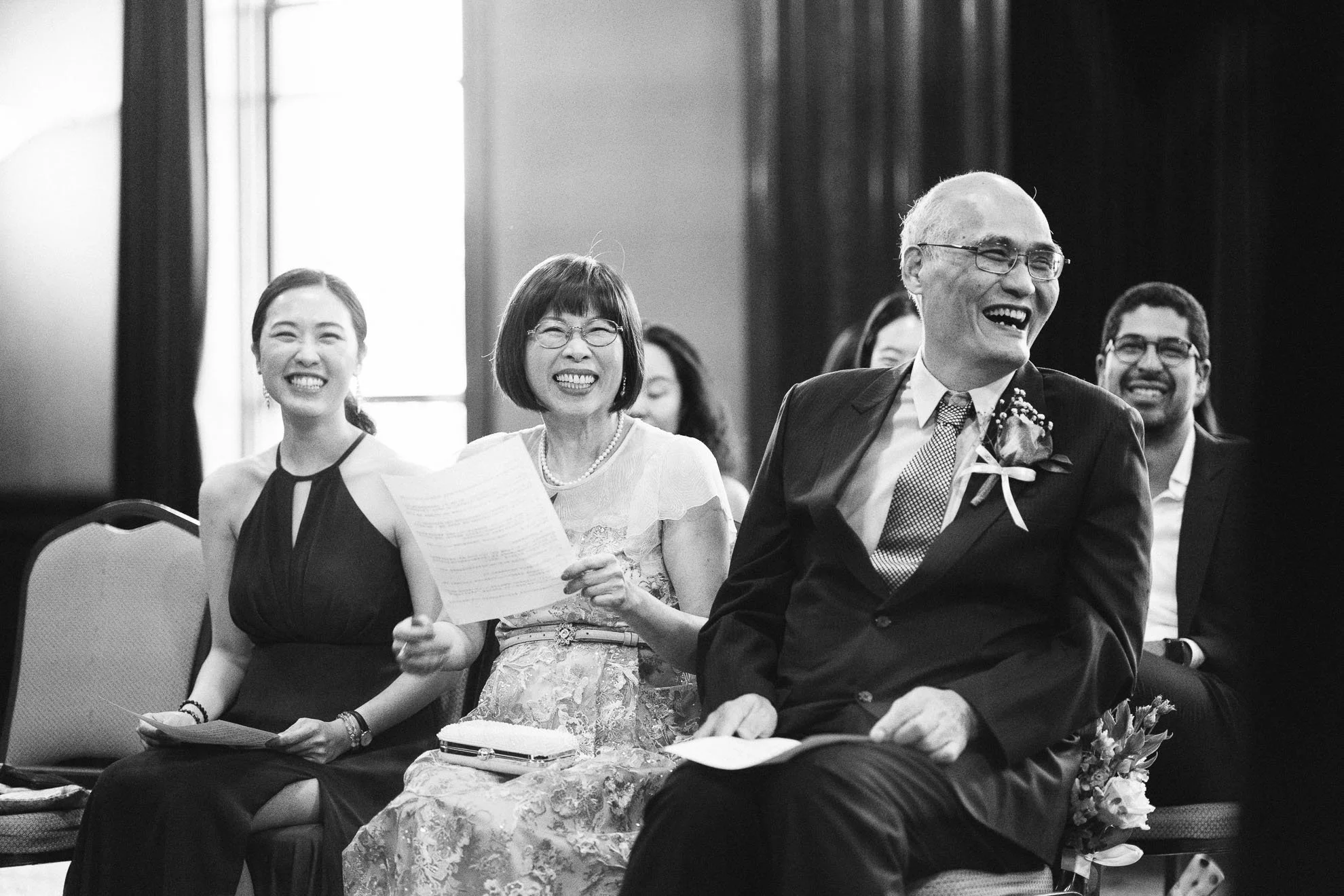 Black-and-white photo of people at a formal event, smiling and sitting in rows, with others in the background looking on.