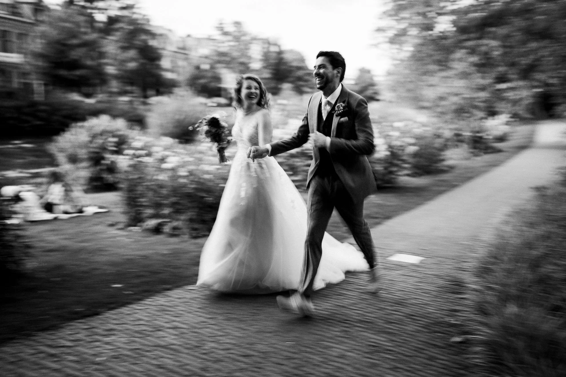 A black and white photo of a newlywed couple running outdoors, holding hands and smiling joyfully, with the bride holding a bouquet and wearing a wedding dress, and the groom in a suit.