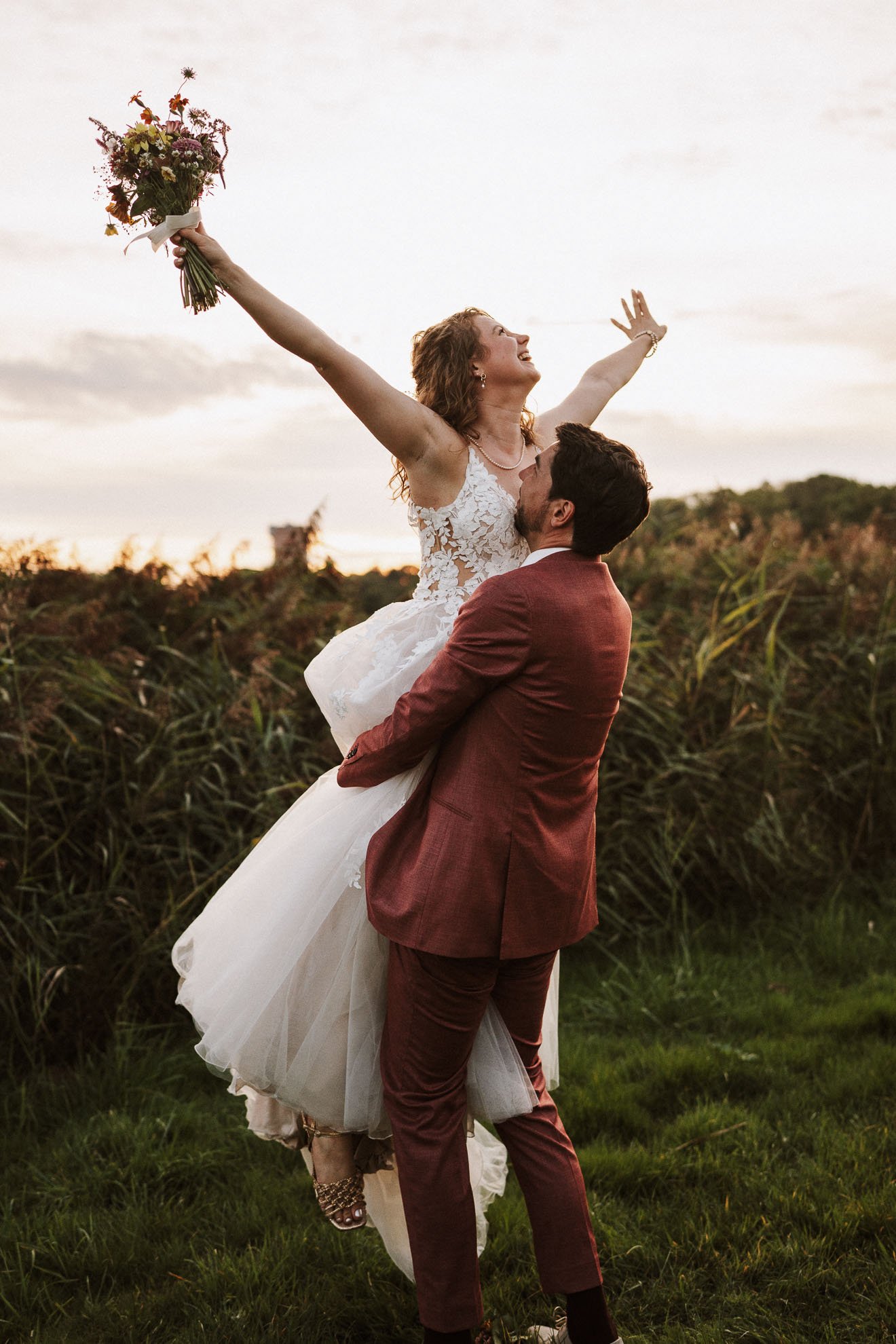 A woman in a white lace wedding dress is being lifted by a man in a reddish-brown suit, holding a bouquet of flowers and celebrating outdoors during sunset.