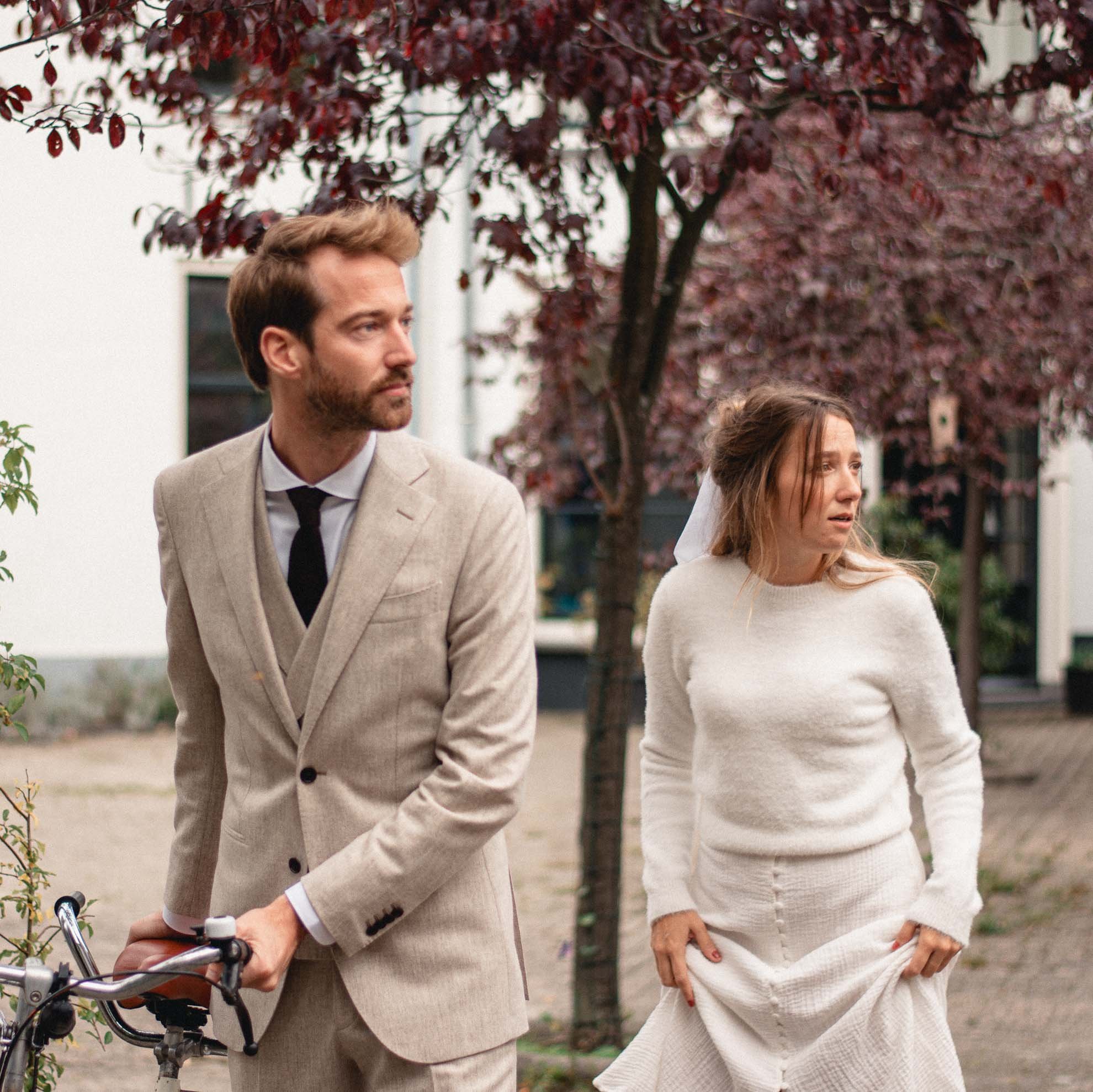 A man and woman walking outdoors near a red-leaved tree, with the man holding a bicycle handlebar.