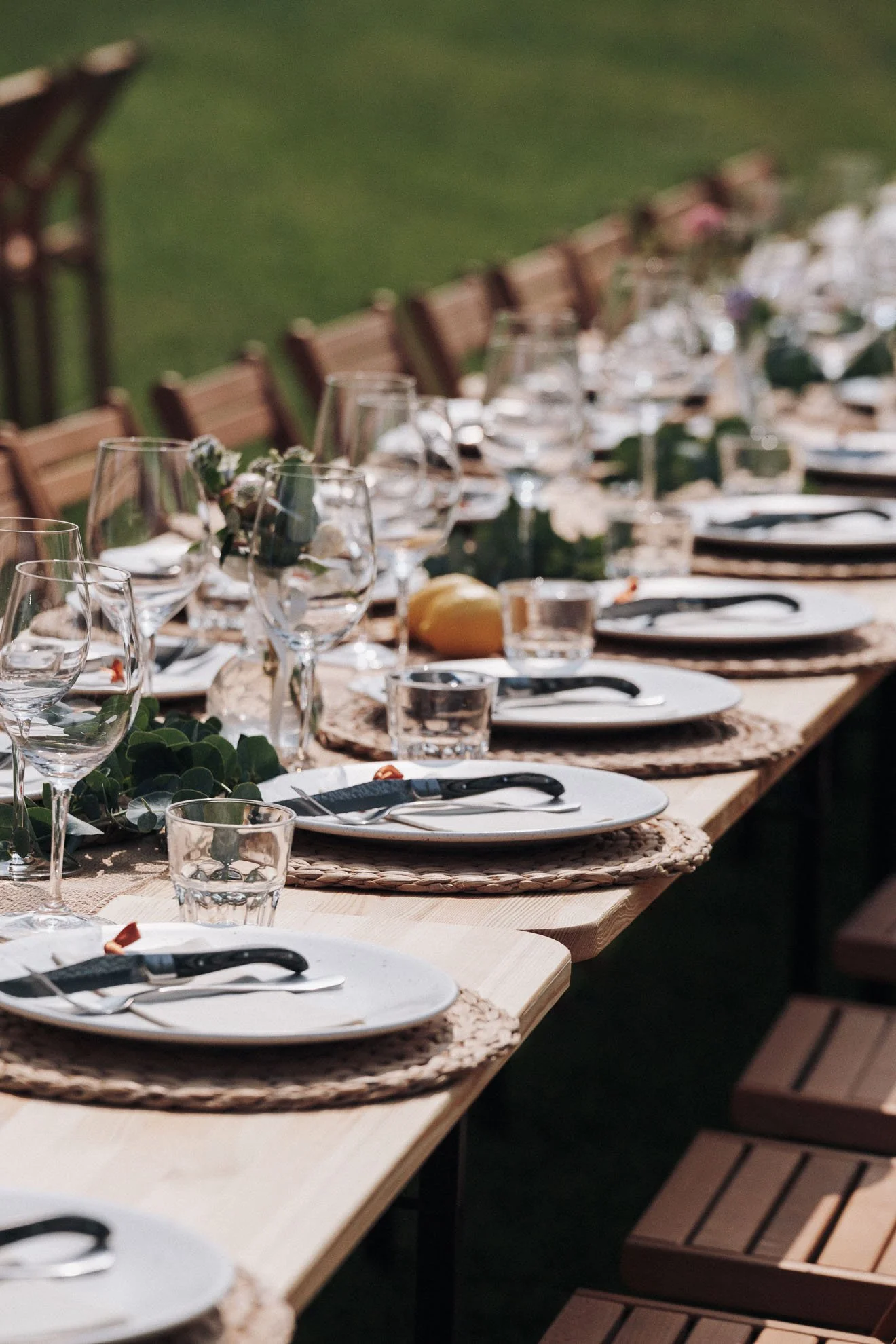 A long outdoor dining table set for a meal with wine glasses, plates, cutlery, and a table runner. The table is decorated with greenery and surrounded by wooden chairs. The background shows a green outdoor area.
