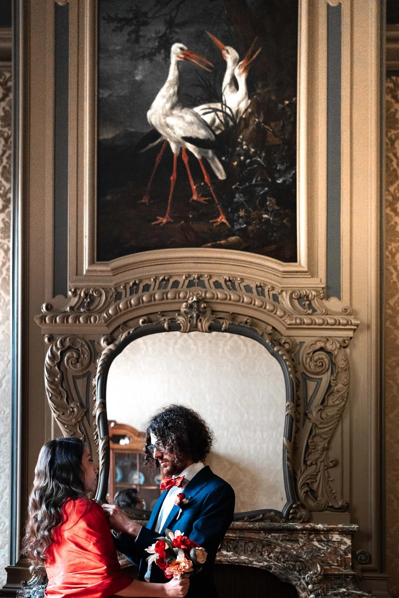 A couple getting married, standing in front of a large ornate mirror, with a painting of two storks above it.
