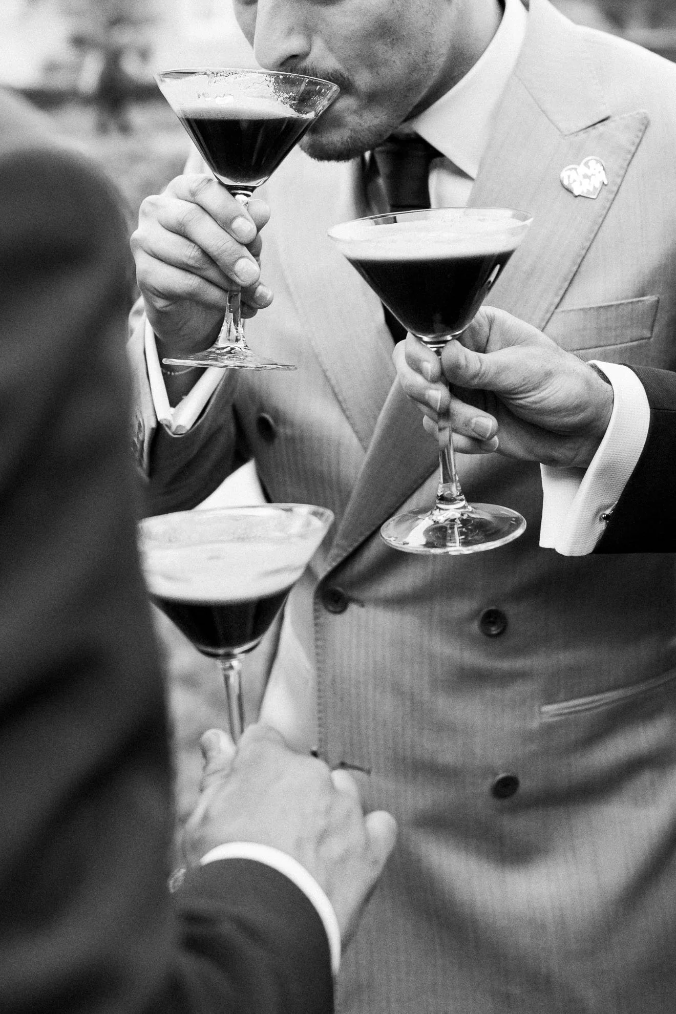 A black-and-white photo of a man in a suit and tie holding two cocktail glasses, appearing to taste or smell the drinks.