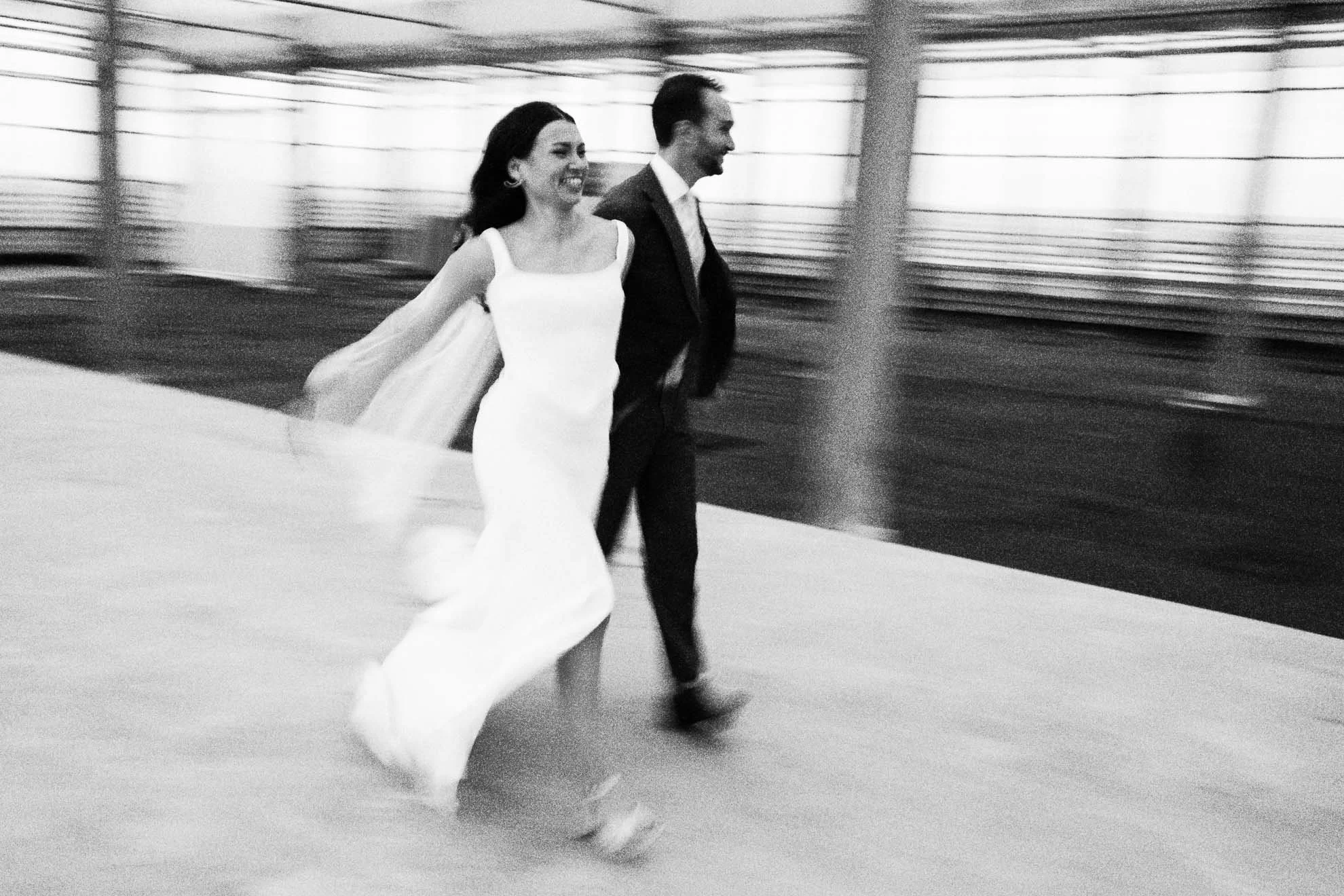 Black and white photo of a smiling woman in a white dress and a man in a suit walking together, blurred background indicating motion.