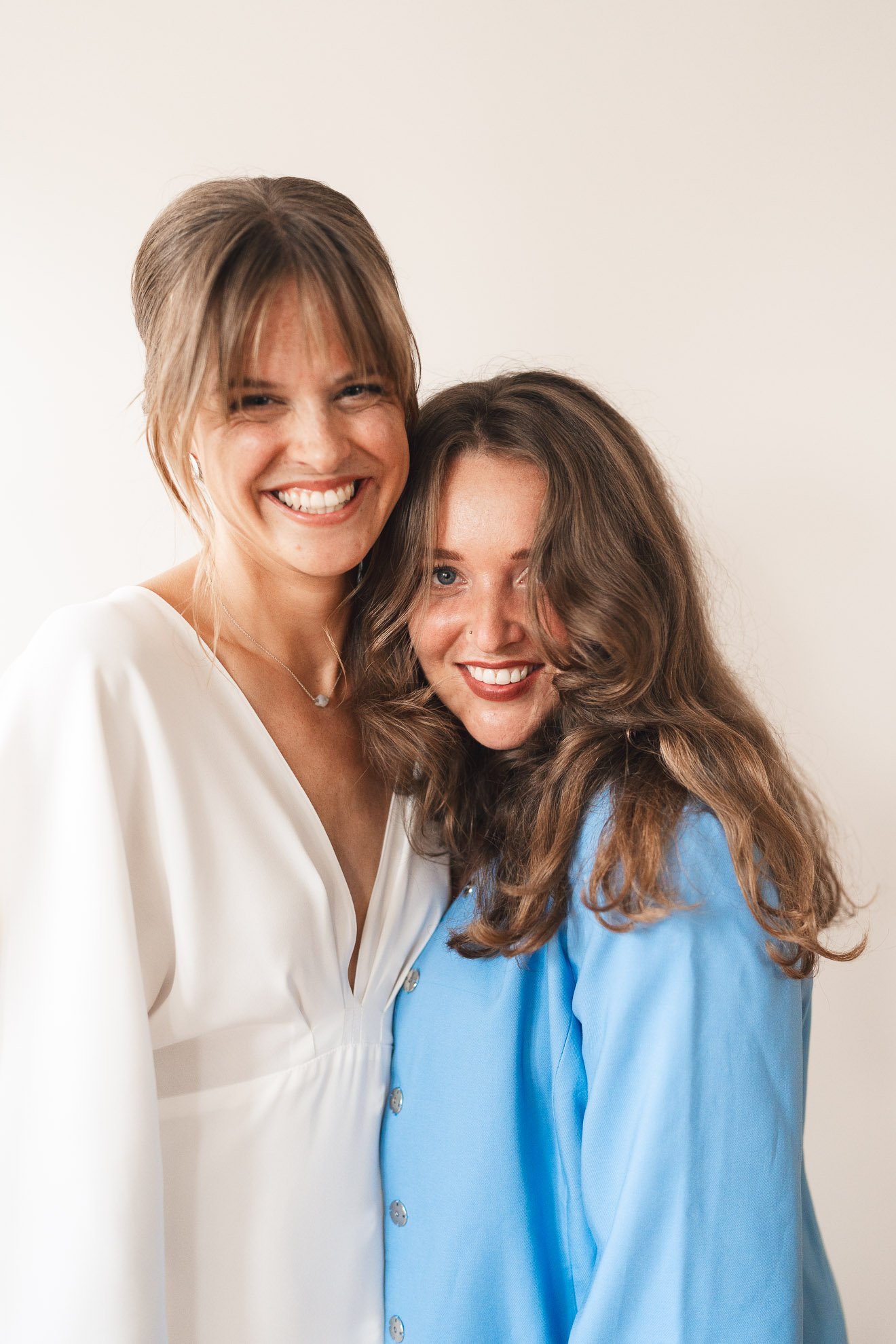 Two women smiling and hugging each other, one with short brown hair and the other with long, curly brown hair, standing against a plain background