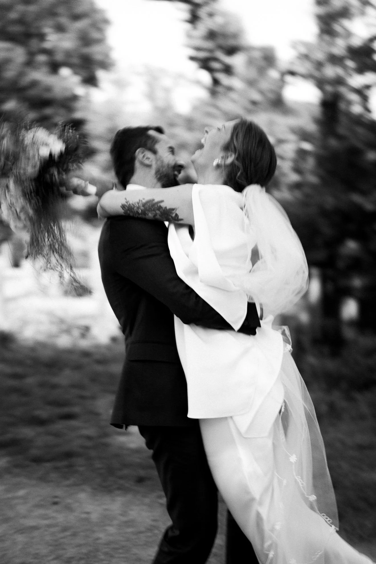 A happy couple in wedding attire embracing outdoors, with the bride holding the groom, both smiling and looking at each other.