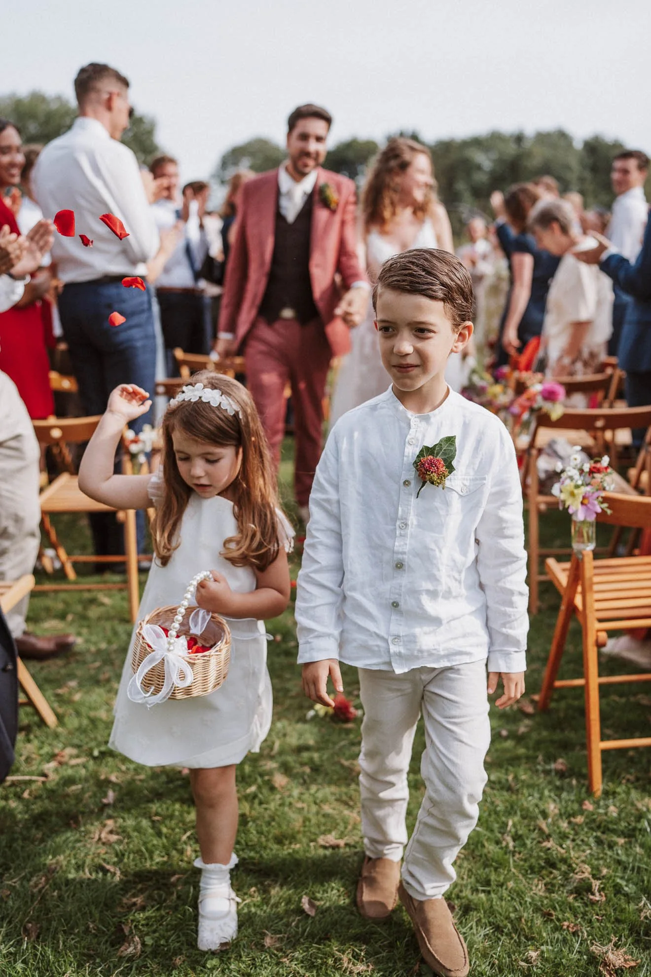 Children walking down an aisle at a wedding, with guests clapping and rose petals in the air. The children are dressed in white, and wedding chairs decorated with flowers are visible.
