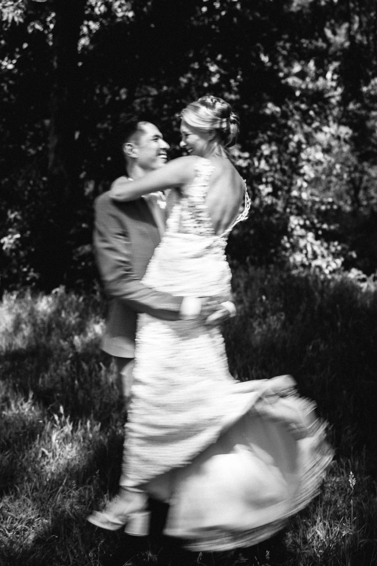 A black-and-white photo of a couple dancing outdoors, with trees in the background. The woman is wearing a long dress, and the man is dressed in a suit. They are smiling and looking at each other, enjoying the moment.