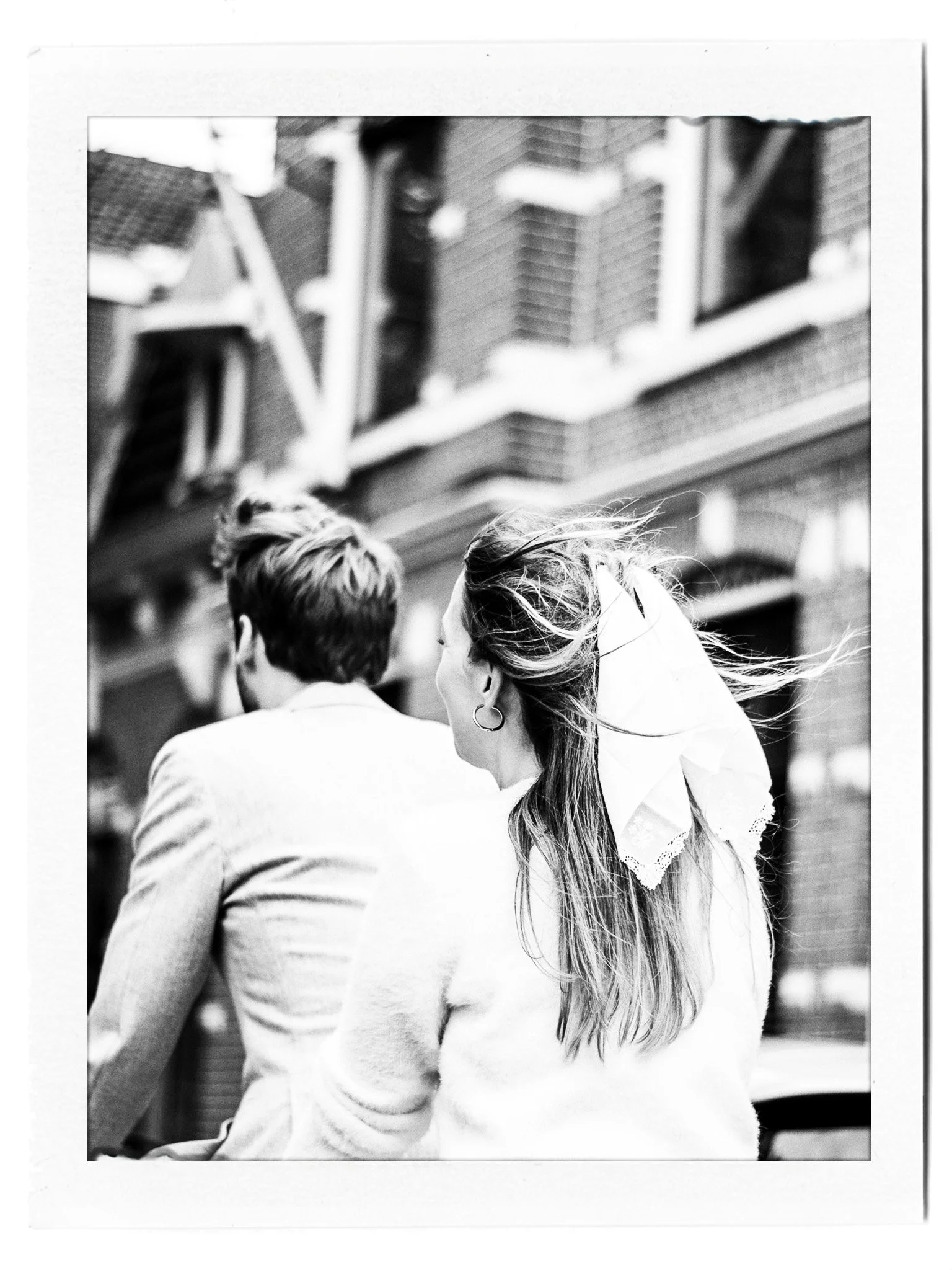 Zwart-wit foto van een vrouw met lange haar en oorbellen, die naast een man met kort haar en een blazer zit, buiten in een stad met een gevel met bakstenen in de achtergrond.