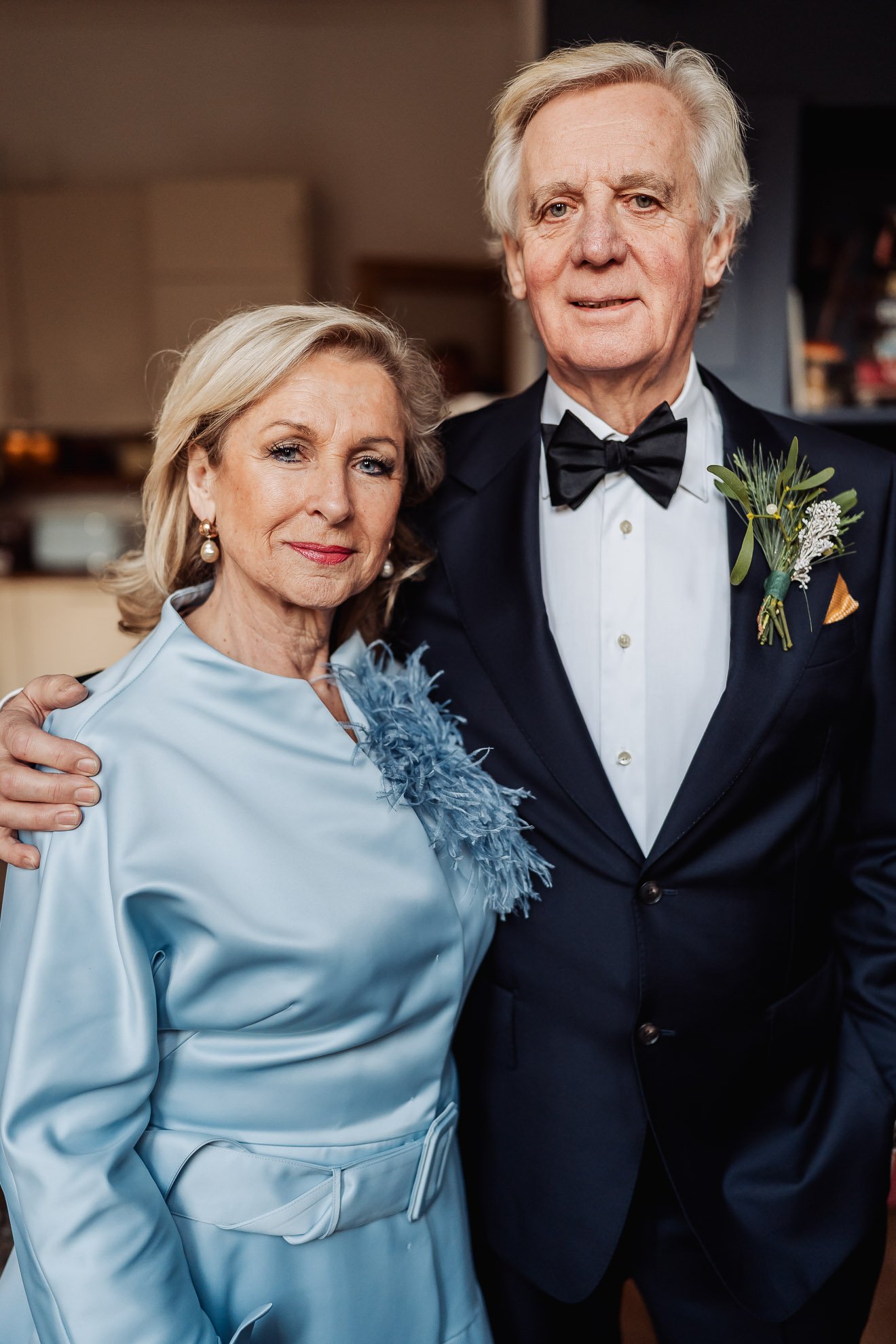 An elderly couple dressed elegantly, standing close together indoors. The woman is wearing a light blue dress with feather detail on the shoulder, pearl earrings, and soft makeup. The man is in a black tuxedo with a white shirt, black bow tie, and bo