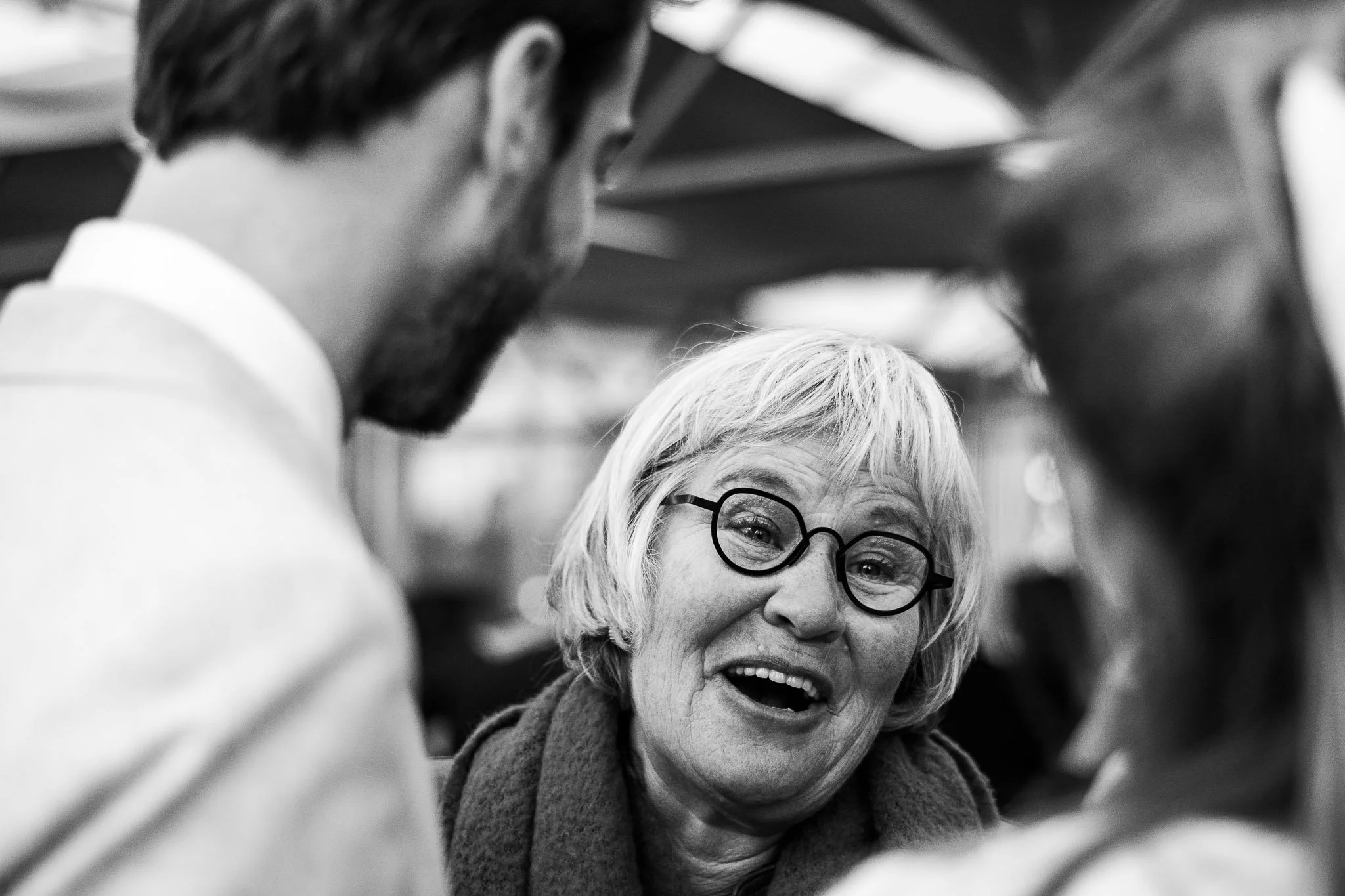 Een glimlachende oudere vrouw met ronde bril, omringd door twee personen en een paraplu, in zwart-wit.