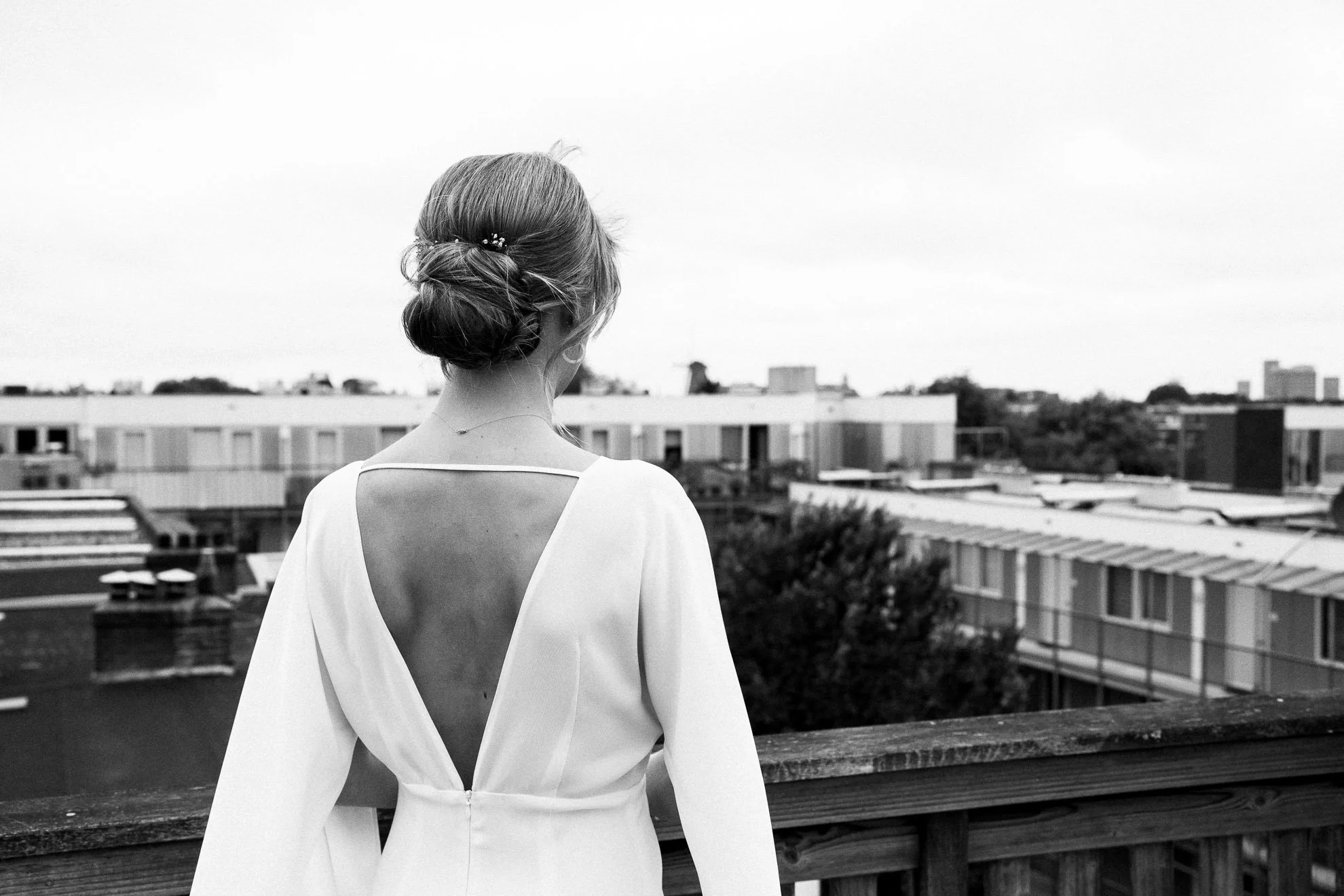 A woman with her hair styled in a low bun, wearing a white dress with an open back, standing on a rooftop or balcony overlooking a cityscape in black and white.