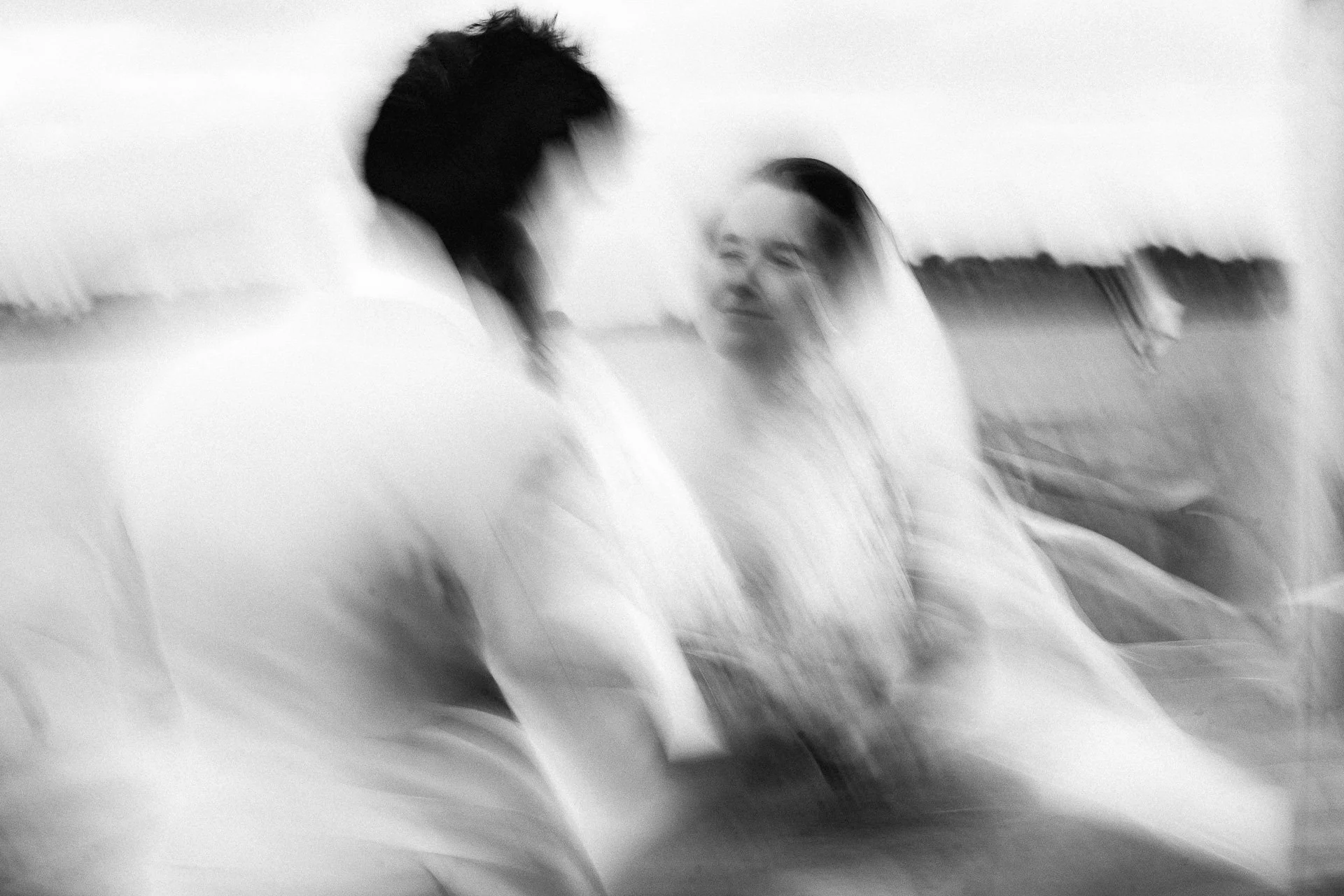 Twee mensen op een romantisch moment op het strand, gekleed in witte kleding, met een derde persoon op de achtergrond, in zwart-wit