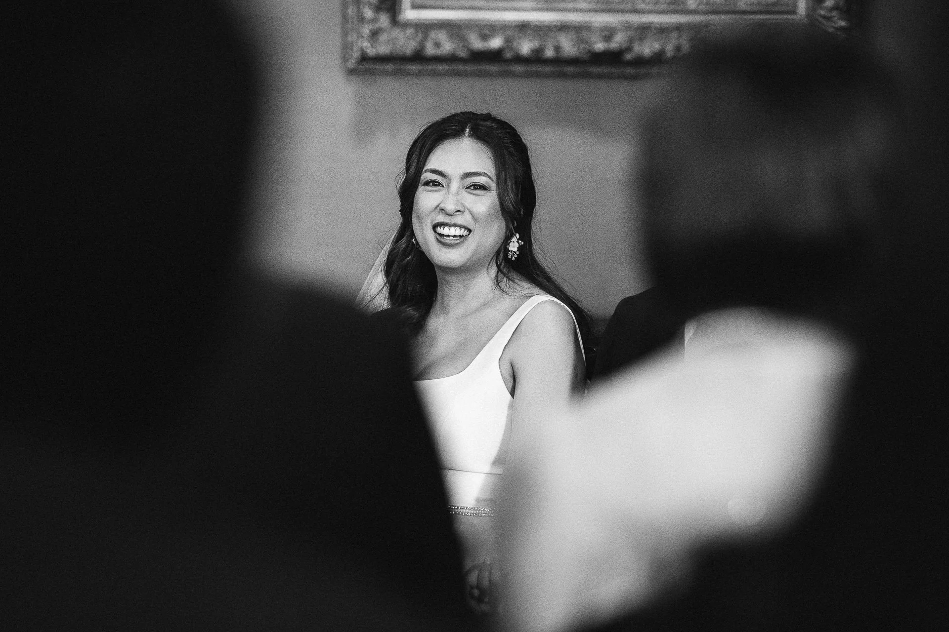 Een gelukkige vrouw in een witte jurk met oorbellen, lachend binnen terwijl haar fotoshoot of evenement plaatsvindt, omringd door mensen.