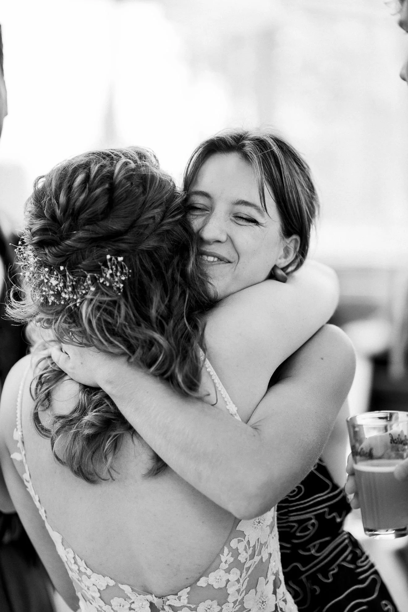 Two women warmly embrace at a celebration, with one woman smiling with eyes closed and holding a glass of beer. The woman facing away has styled hair with braids and floral accessories, and is wearing a dress with floral lace details.