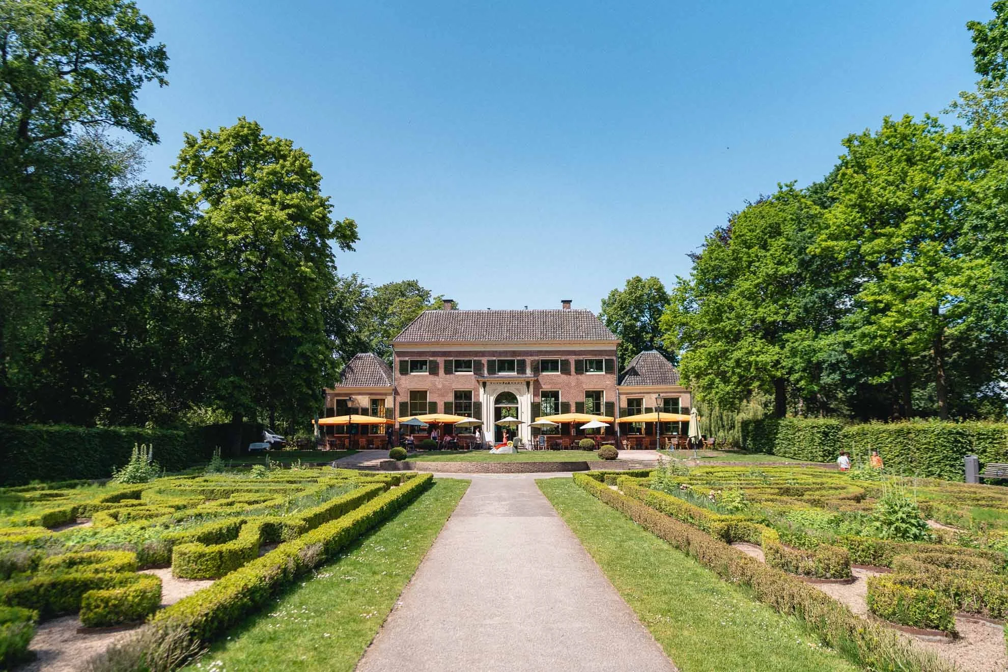 A large, elegant brick mansion with a tiled roof, set amidst lush green trees and a well-manicured garden with a pathway leading to the entrance, outdoor seating with umbrellas, and blue sky overhead.