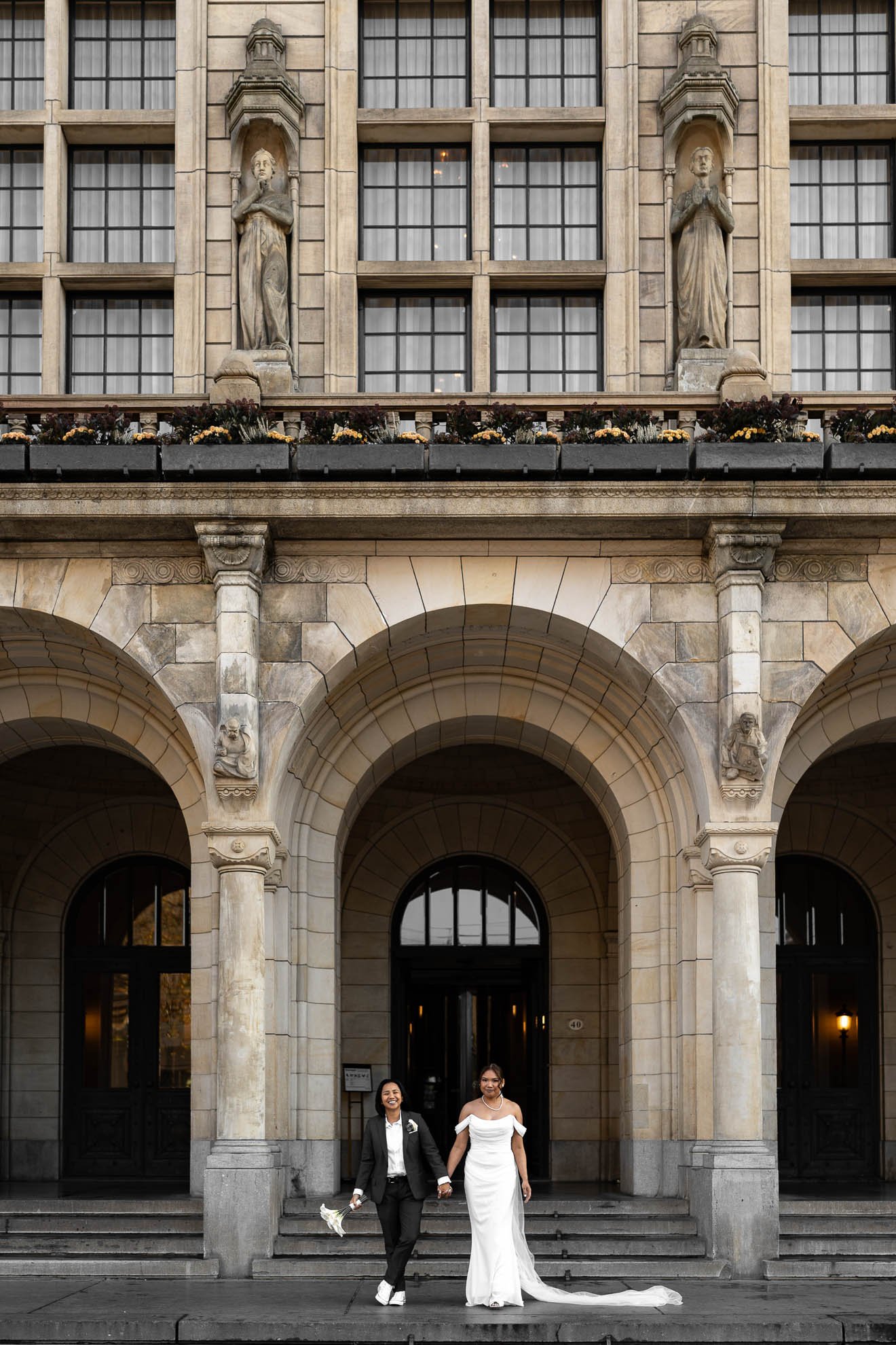 Two women, one in a black suit holding a bouquet and the other in a white wedding gown, walking out of a historic building with stone arches and statues.