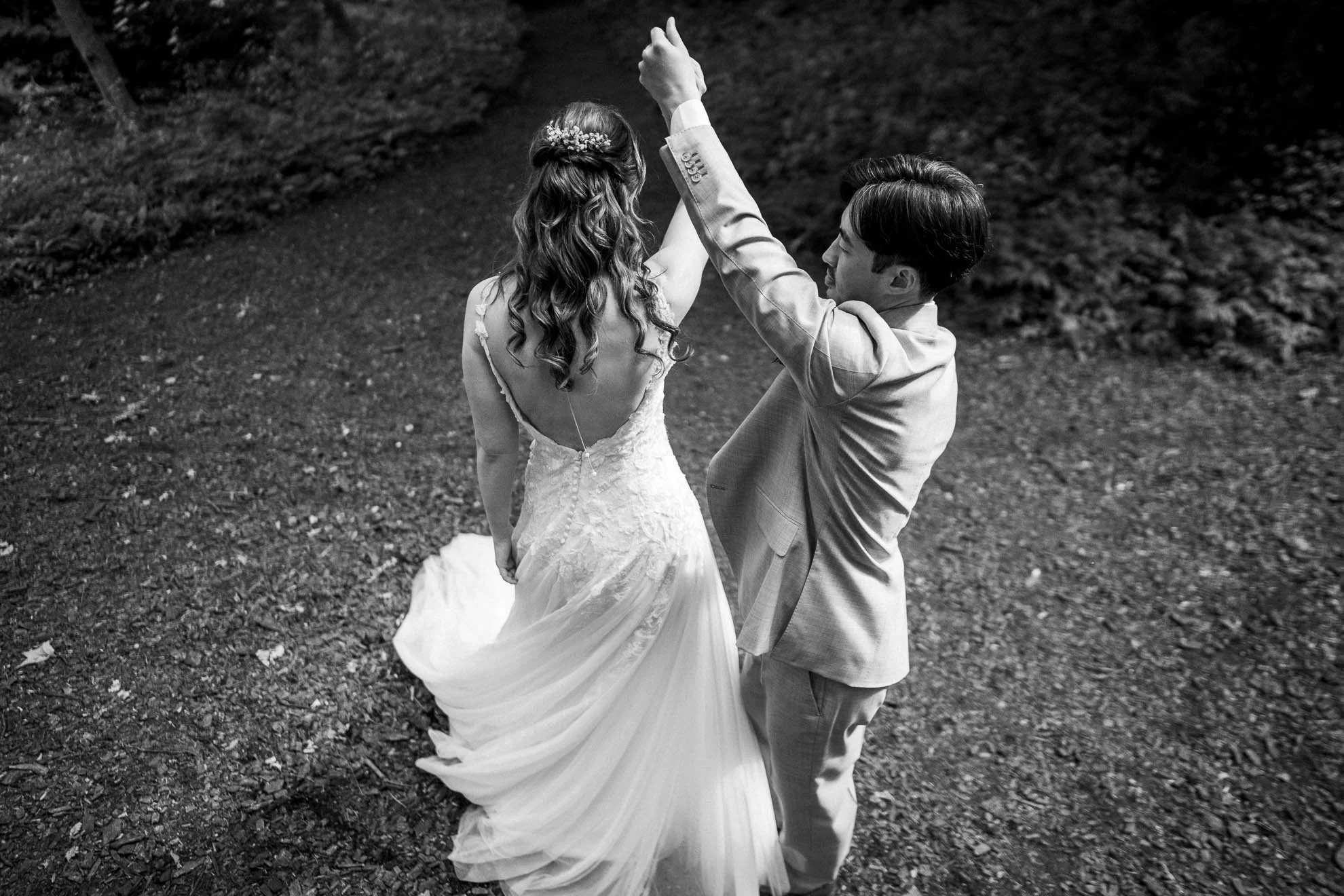 A black-and-white photograph of a bride and groom dancing outdoors on a grass and dirt area surrounded by trees or bushes, with the bride's wedding dress flowing as they dance.