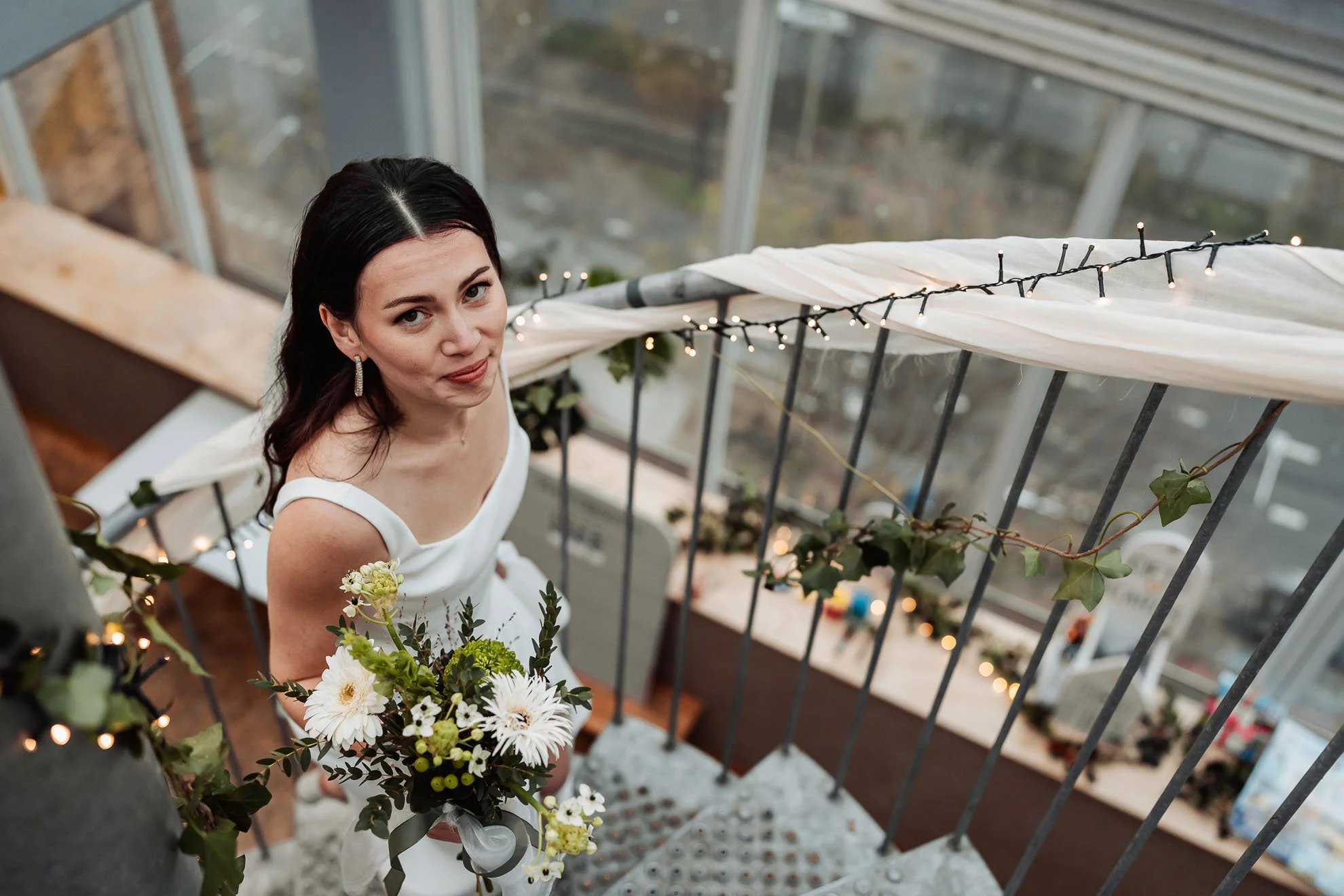 A young woman in a white dress holding a bouquet of white and yellow flowers, standing on a balcony decorated with string lights and greenery, with a cityscape in the background.
